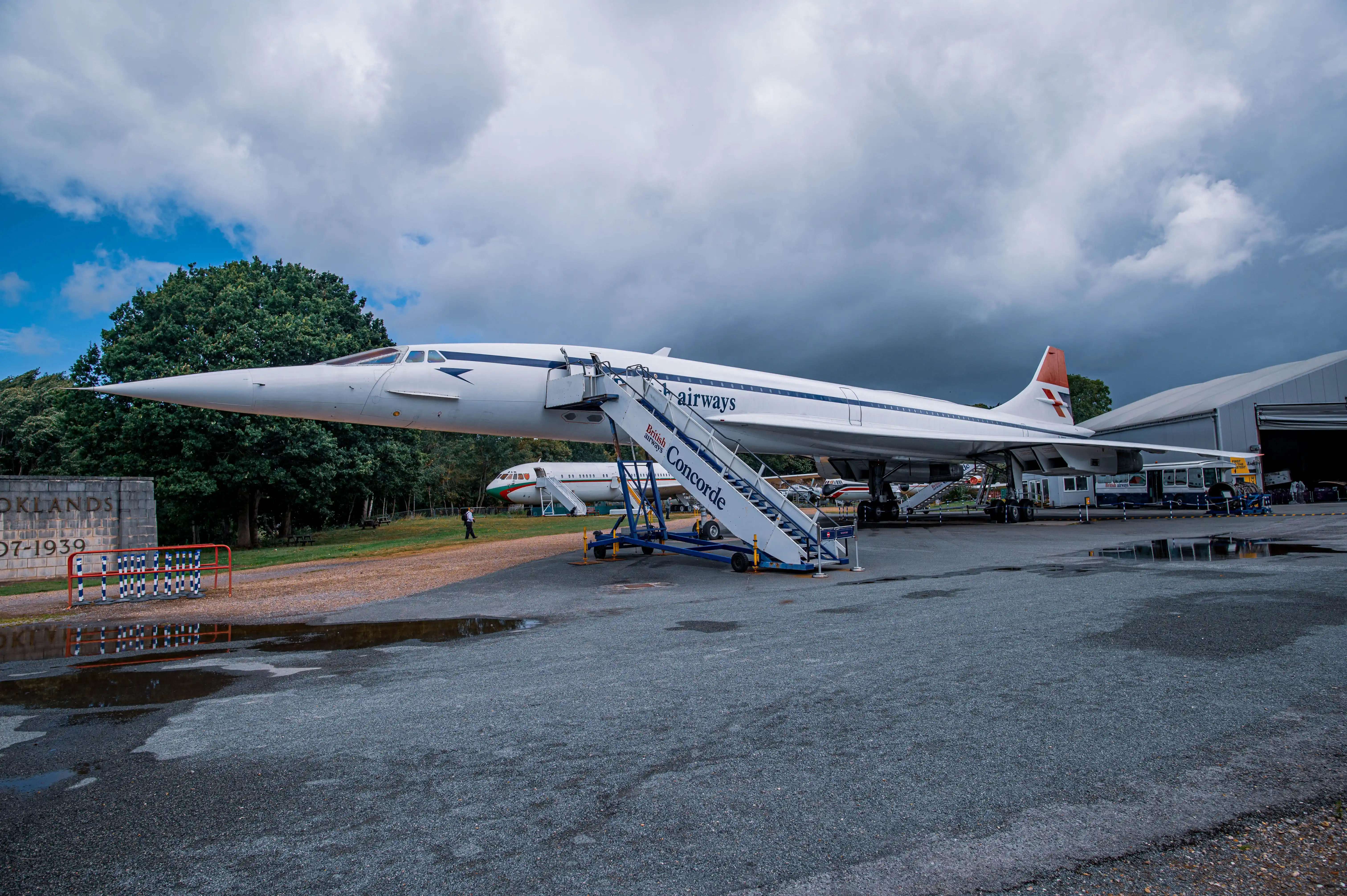 A British Airways Concorde aircraft is parked on display, with a mobile boarding staircase positioned beside it. The aircraft features the iconic white livery with blue stripes. The Brooklands Museum sign is visible in the foreground.