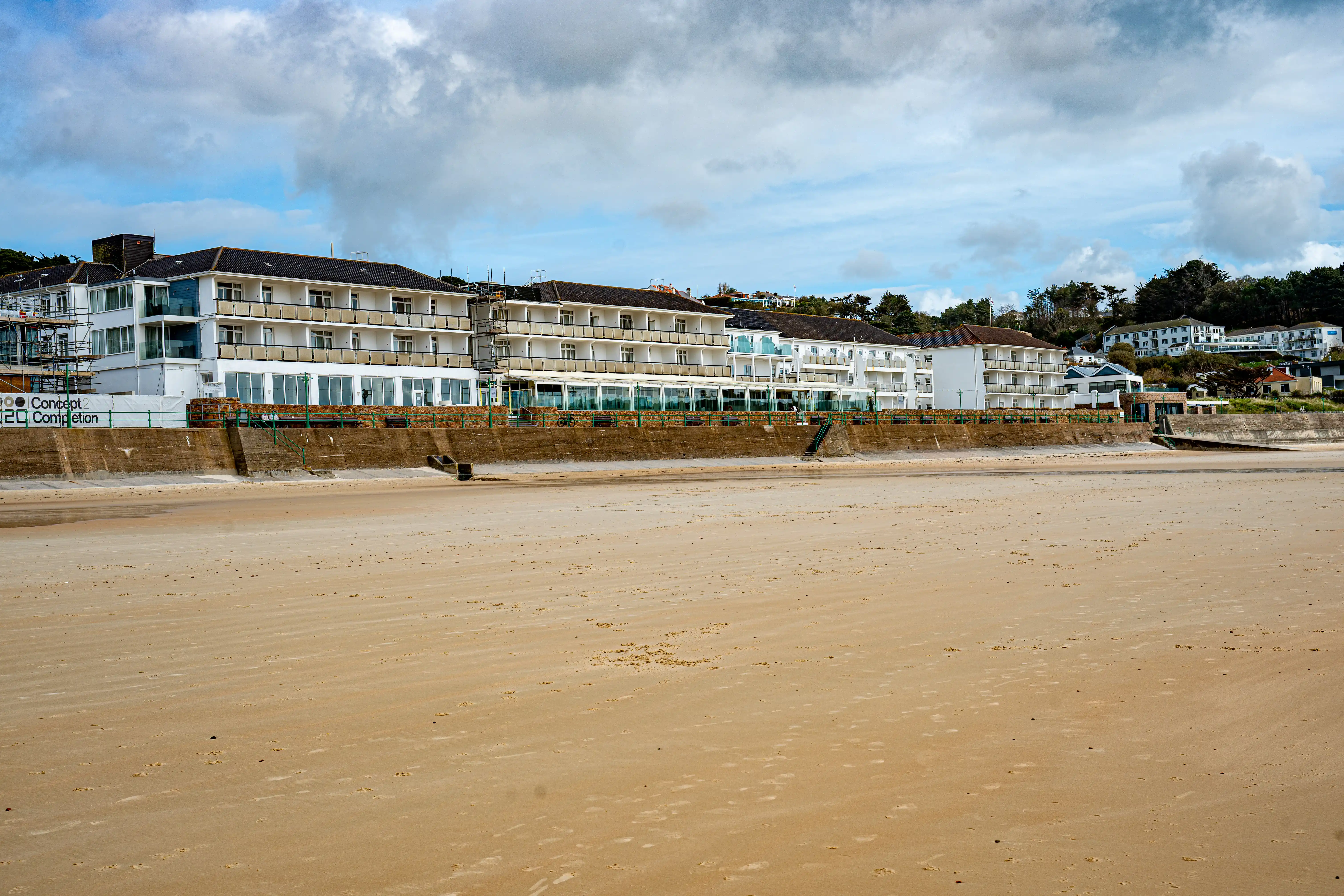 The image depicts a beach scene with a sandy shore in the foreground and a row of modern buildings under construction or renovation in the background