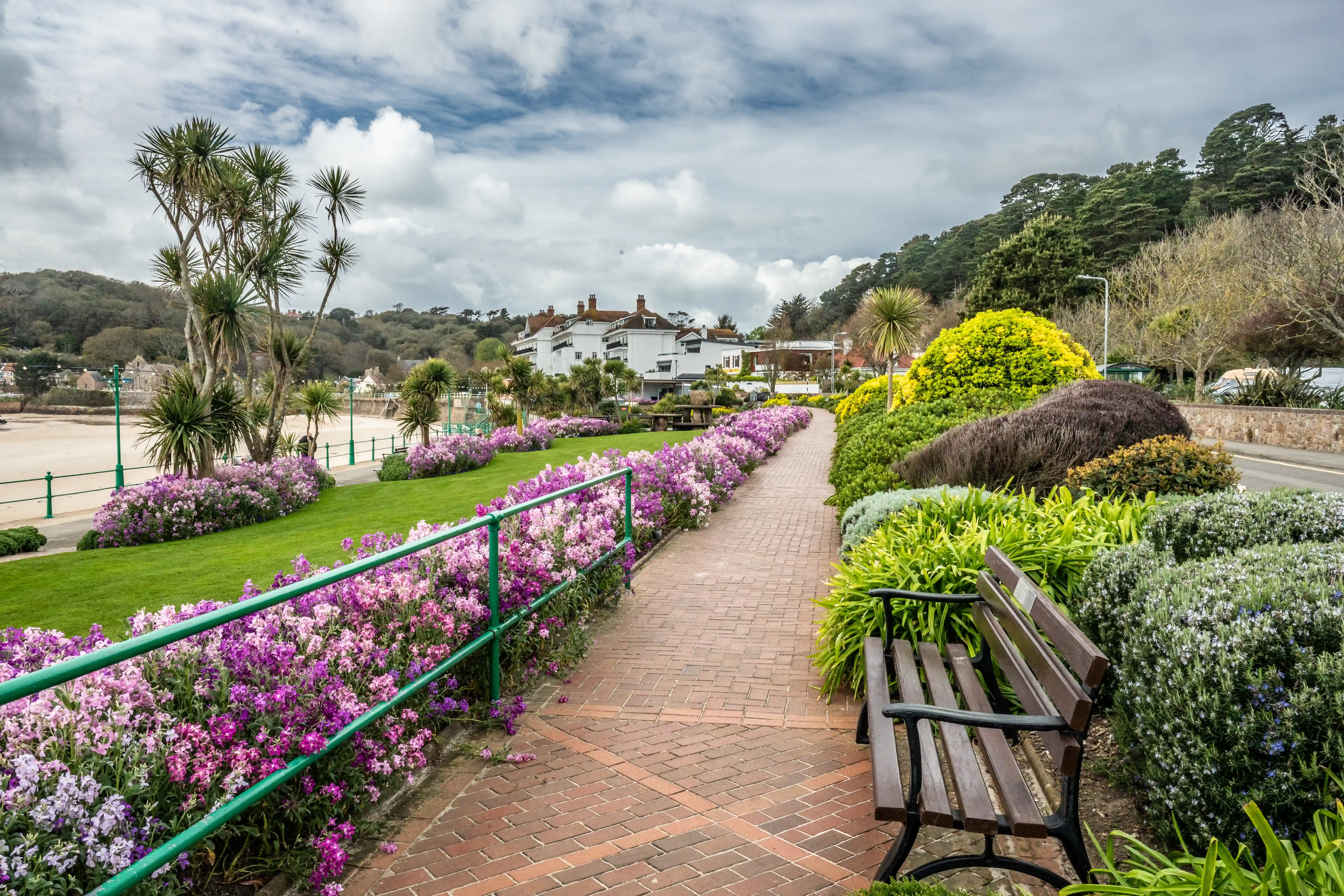 A picturesque paved pathway lined with a vibrant display of purple and pink flowers and manicured greenery, leading towards buildings overlooking a sandy beach. A wooden bench is positioned to the side of the path.