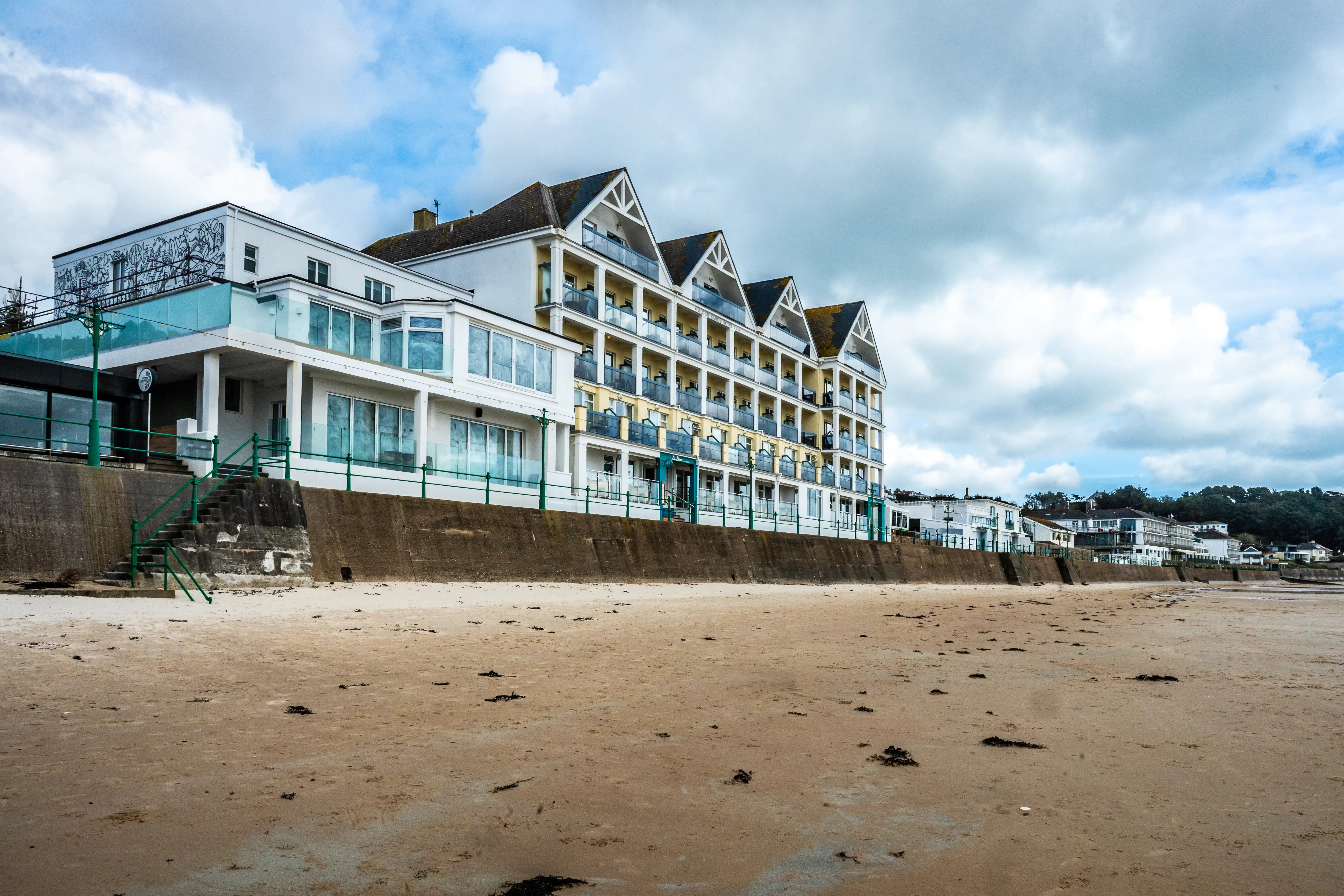 A white and yellow hotel with multiple balconies faces a sandy beach and a concrete sea wall under a cloudy sky.