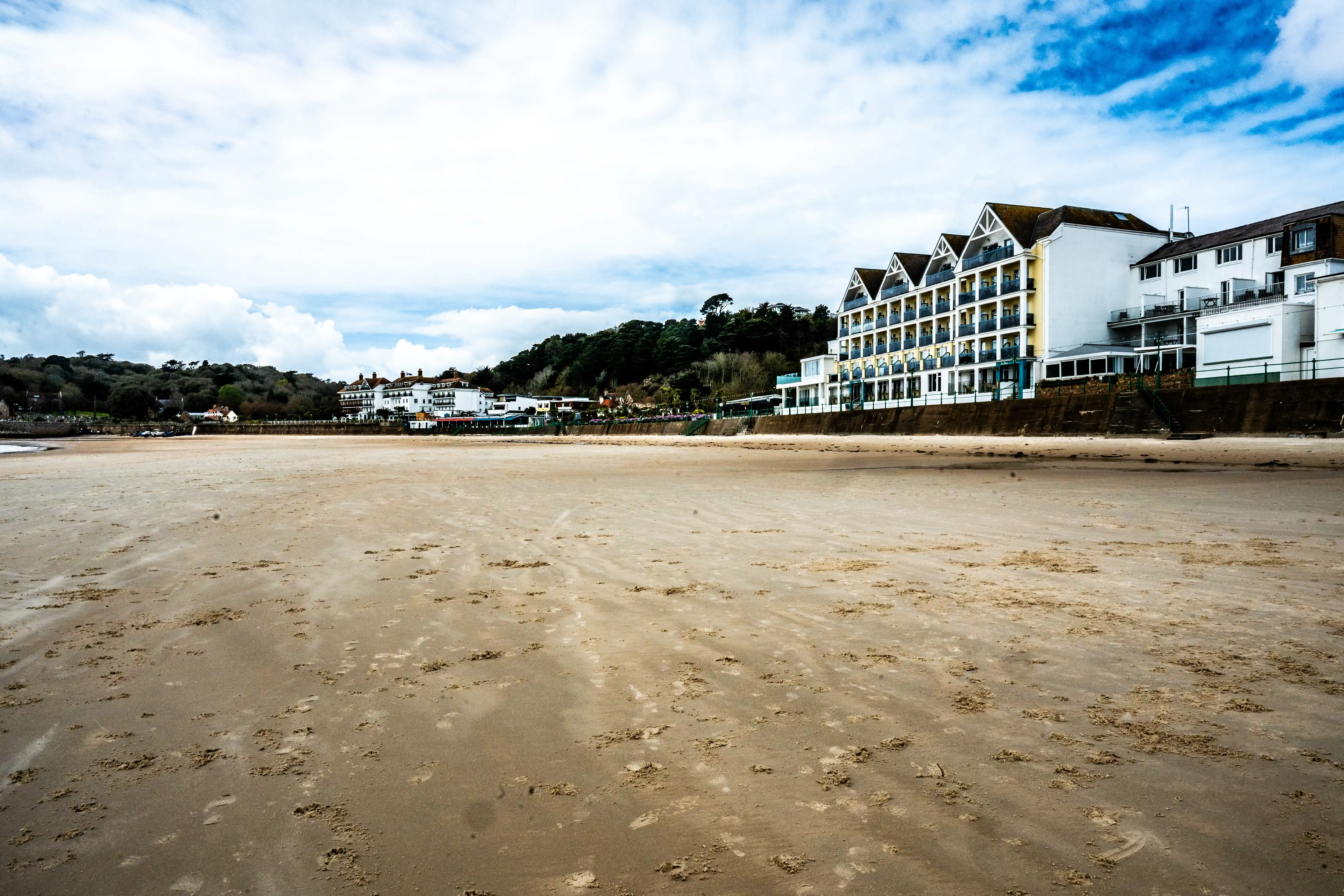 A sandy beach stretches towards a row of buildings, including a prominent hotel with a distinctive triangular roofline, under a partly cloudy sky.
