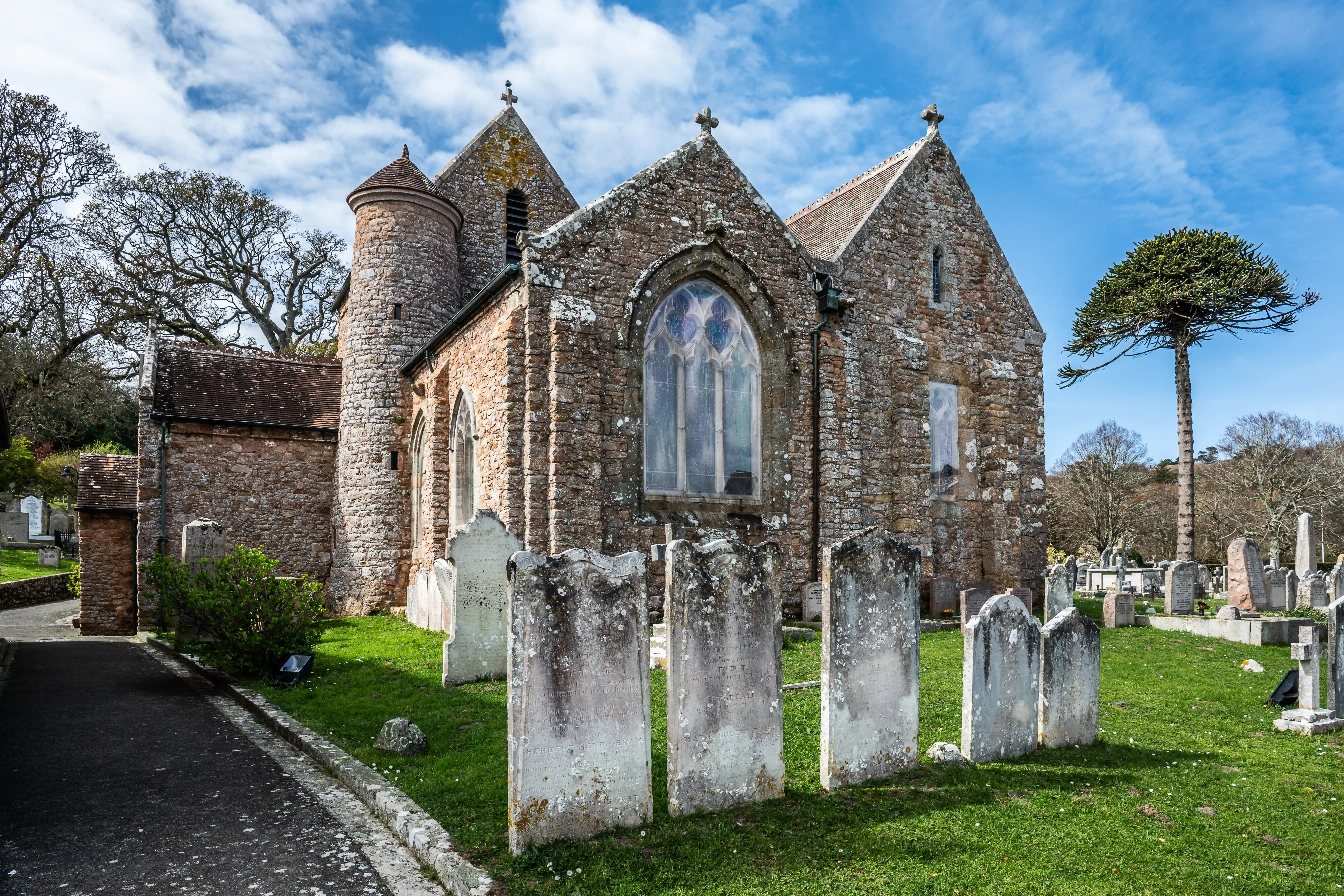  stone church with a round tower and stained-glass windows stands in a graveyard, with weathered gravestones in the foreground and a unique tree in the background under a blue sky with scattered clouds.