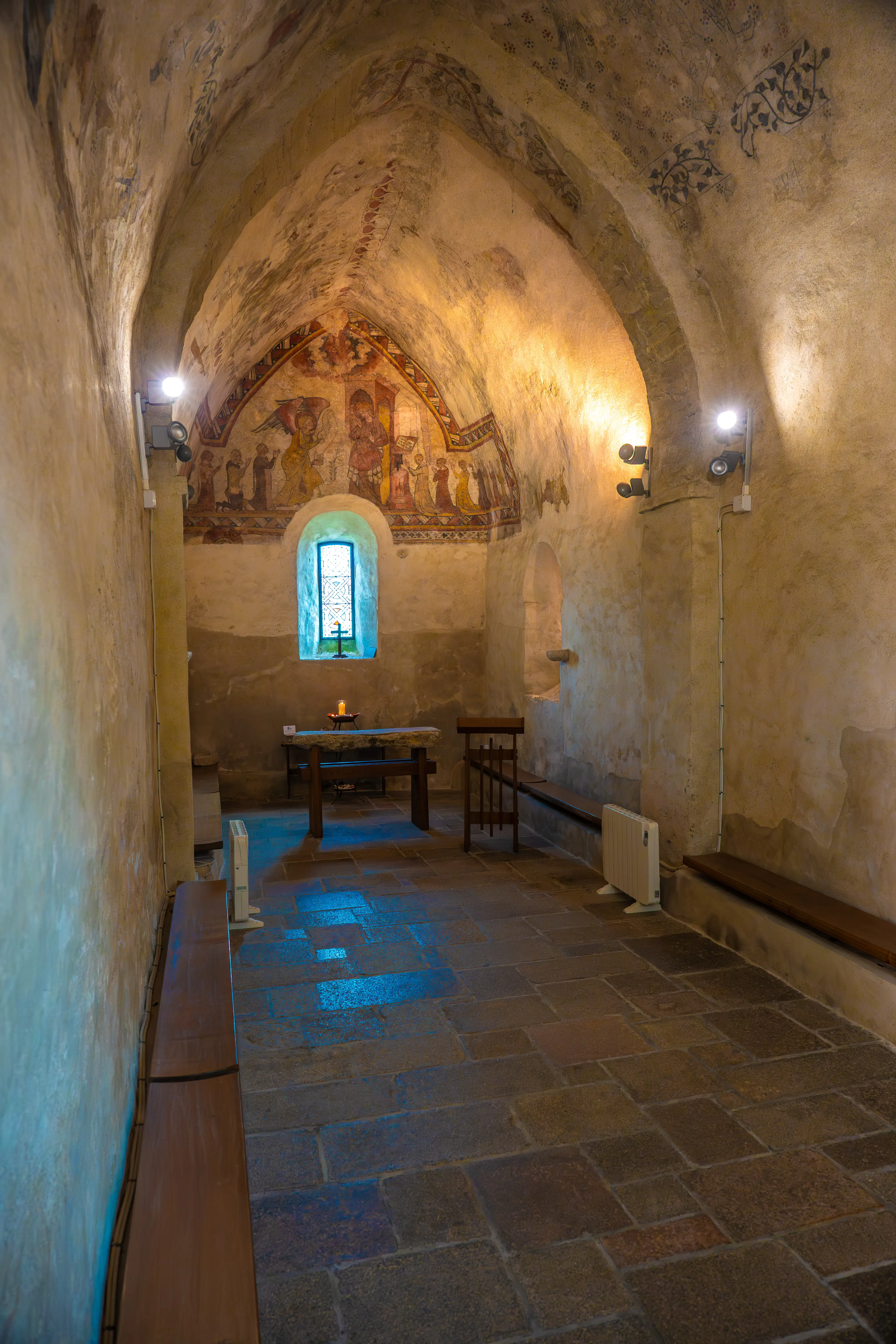 Interior view of an old chapel with arched ceilings painted with frescoes, a stone altar with a lit candle, a stained glass window, and wooden pews.