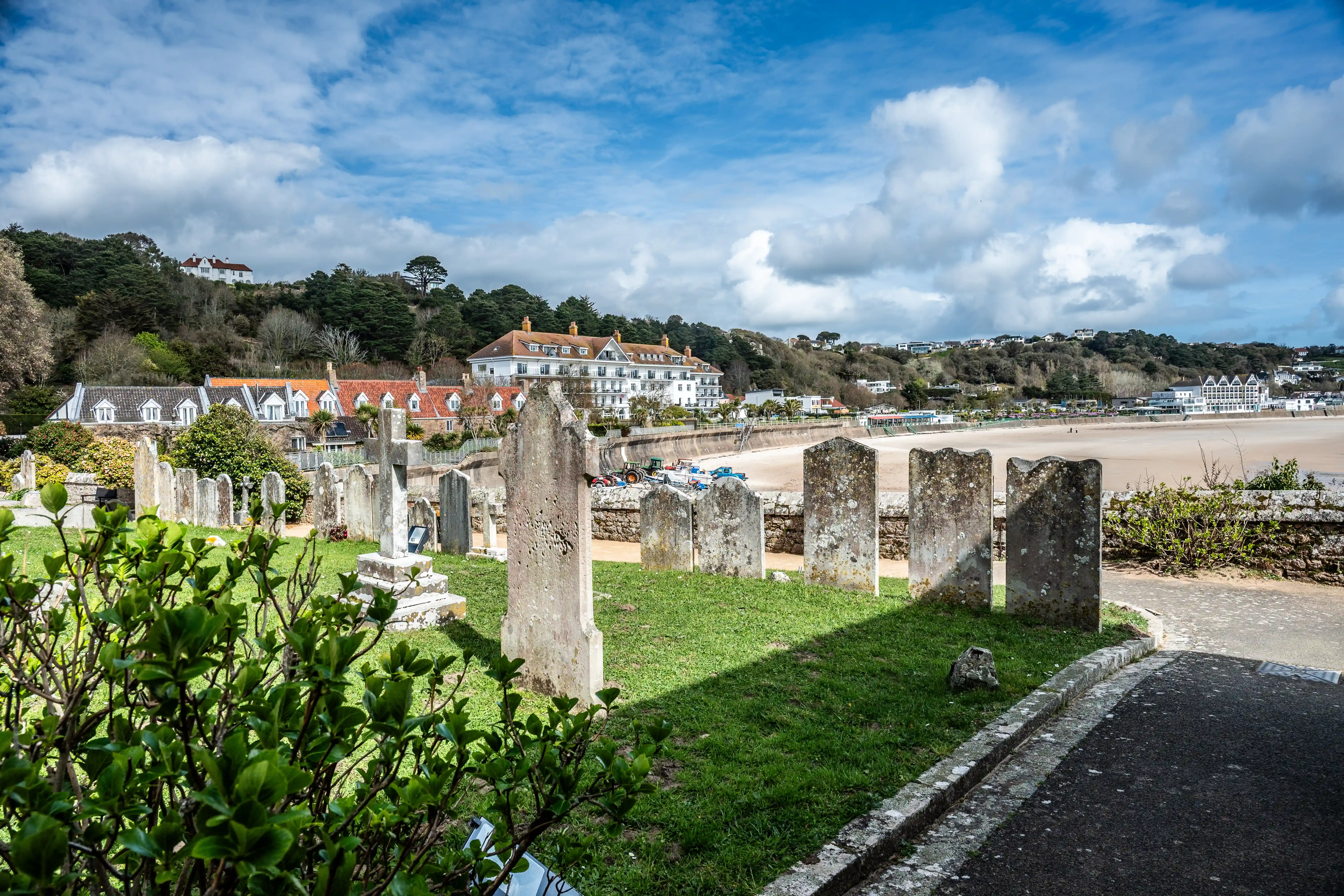 Gravestones in a grassy cemetery overlooking a beach and a town on a sunny day with blue skies and white clouds.