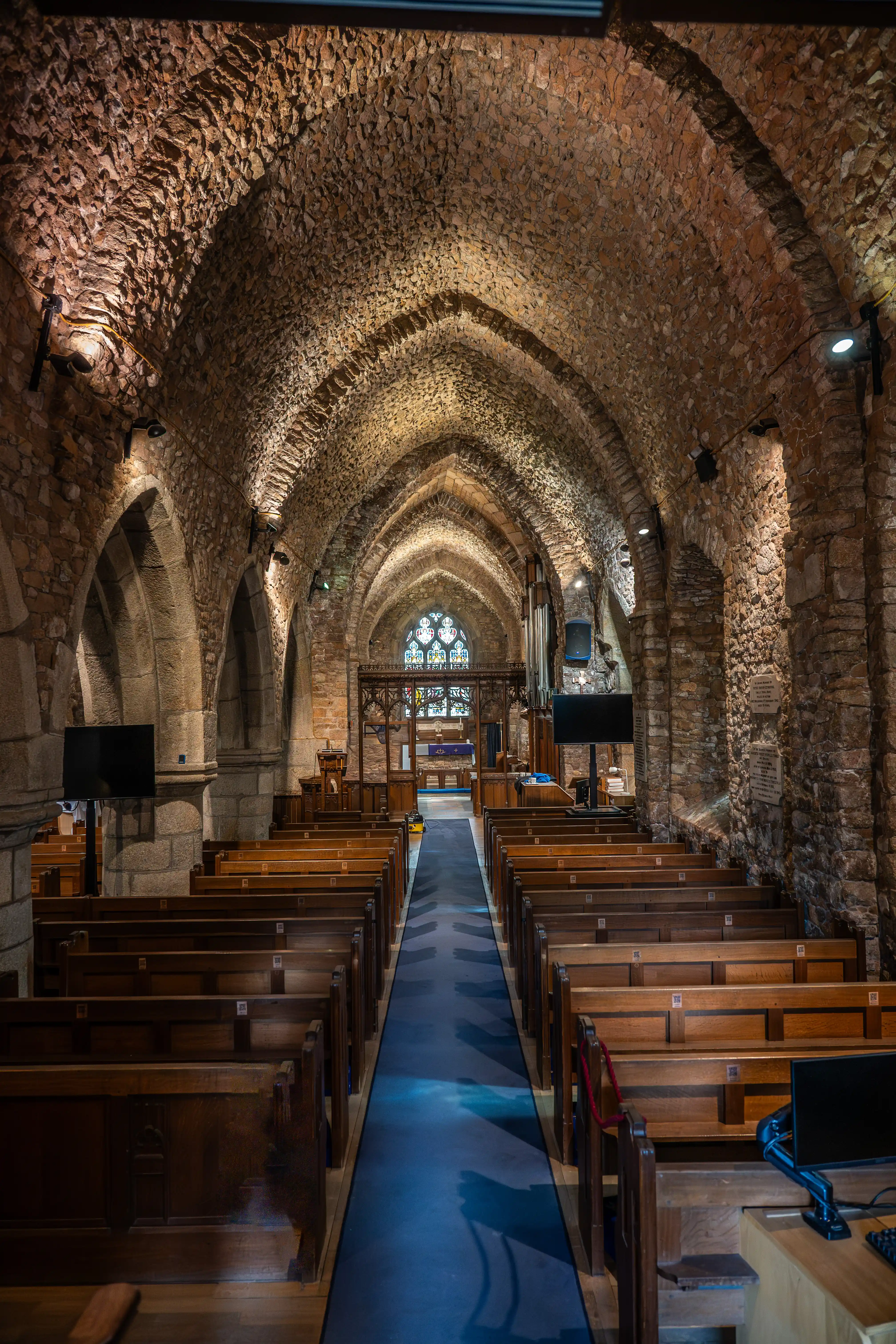 An interior view of a historic church looking down the aisle between rows of wooden pews towards a stained-glass window. The ceiling is made of rough-hewn stone arches, illuminated by spotlights.