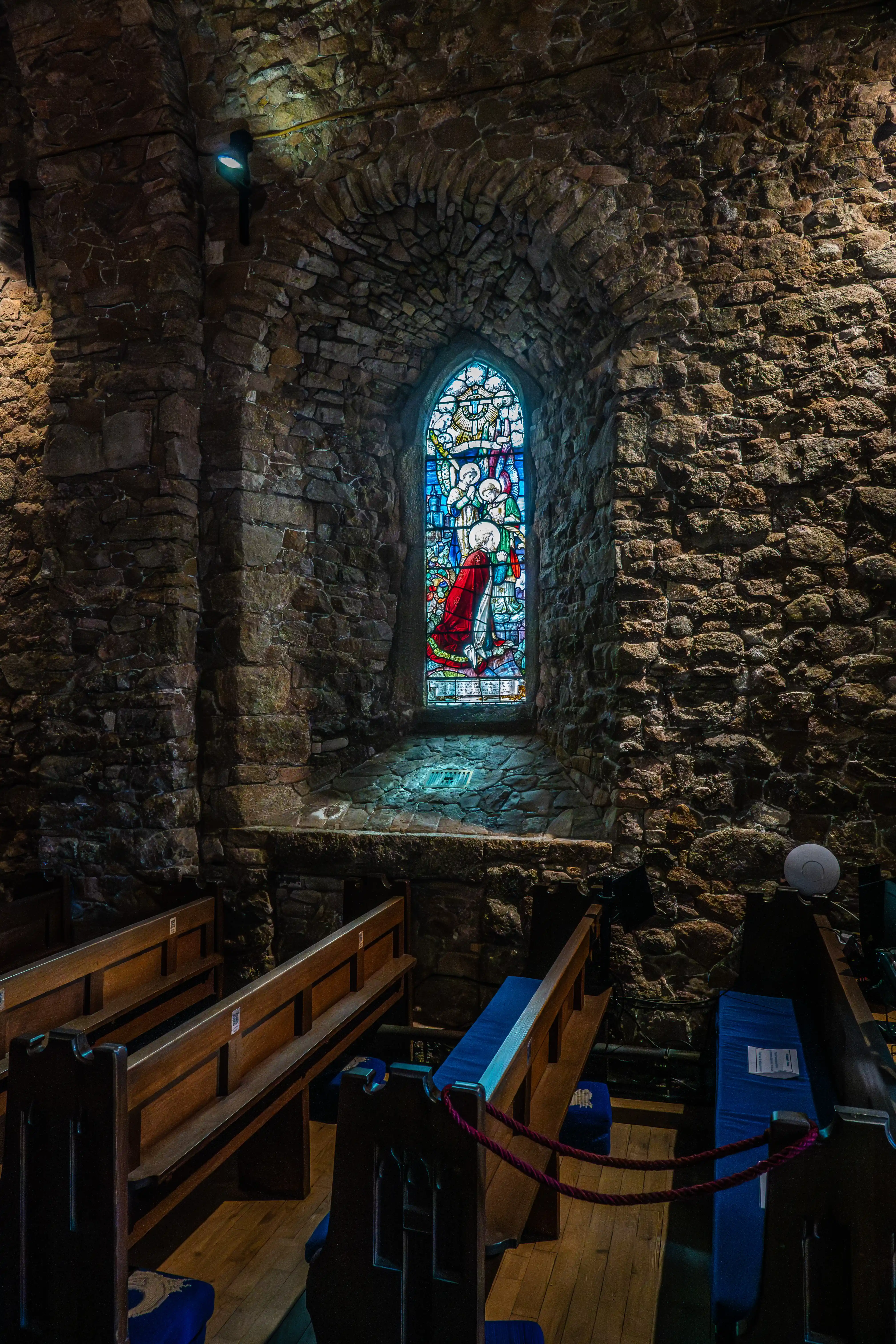  A tall, arched stained-glass window depicting a religious scene with figures and vibrant colours, set within thick, rough-hewn stone walls of a church. Wooden pews are visible in the foreground.