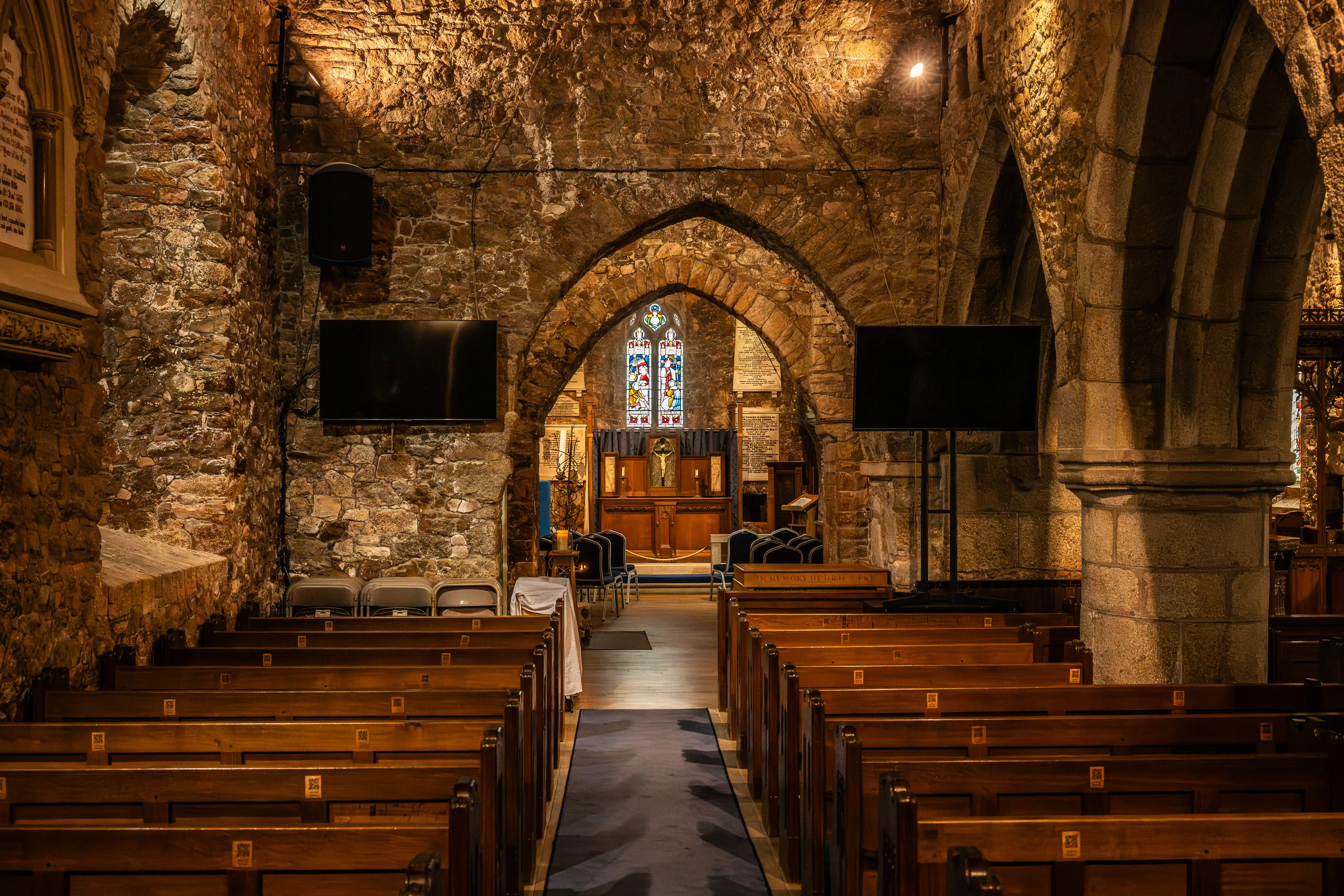 The interior of a church, with wooden pews arranged in rows. The walls are made of rough stone and there are arched stone doorways. At the back of the church, a stained glass window is visible above the altar, which features a crucifix. Two large flat-screen televisions are mounted on the walls.
