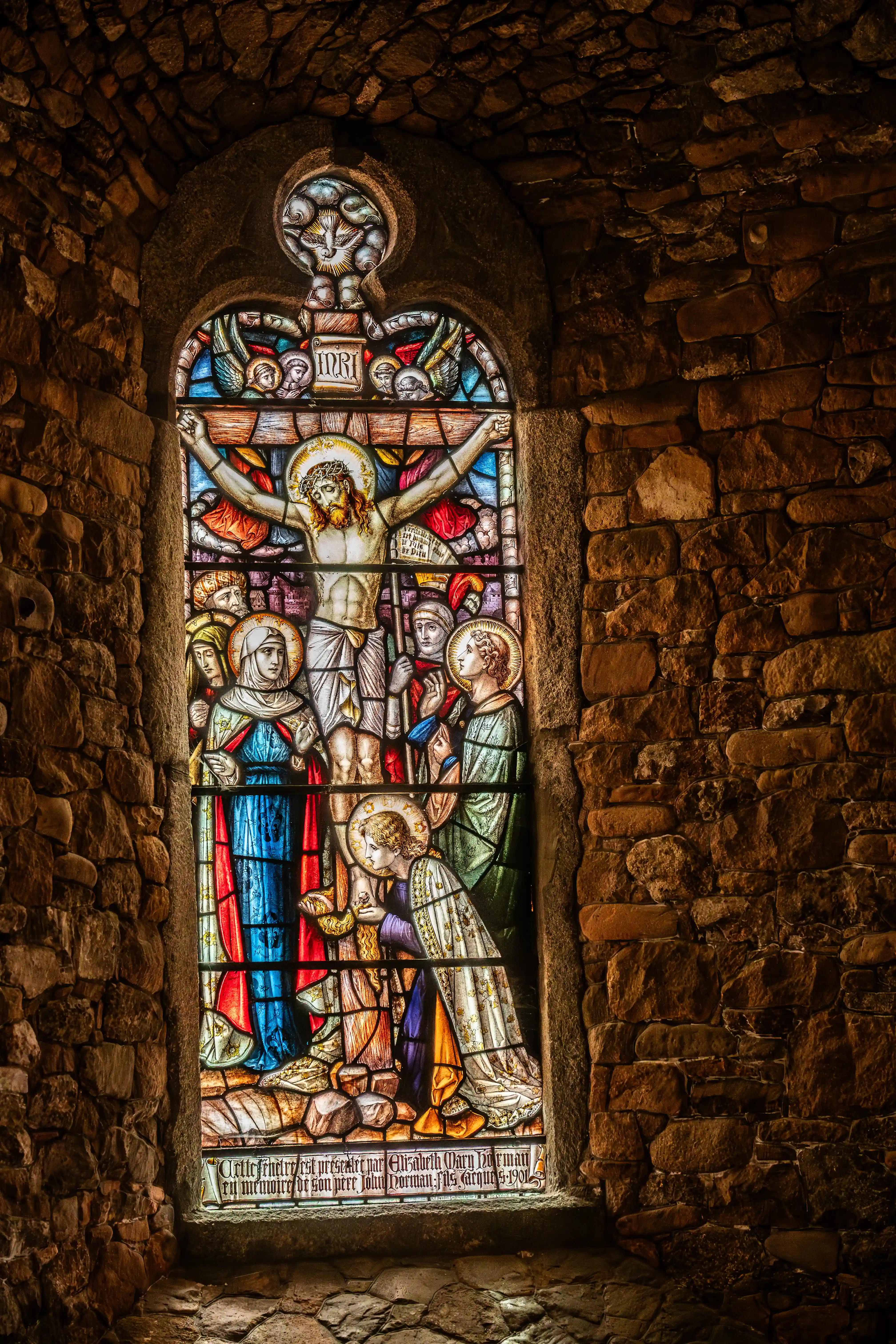 Stained glass window of the Crucifixion, featuring Jesus on the cross, with Mary and other figures below, set within a stone archway.