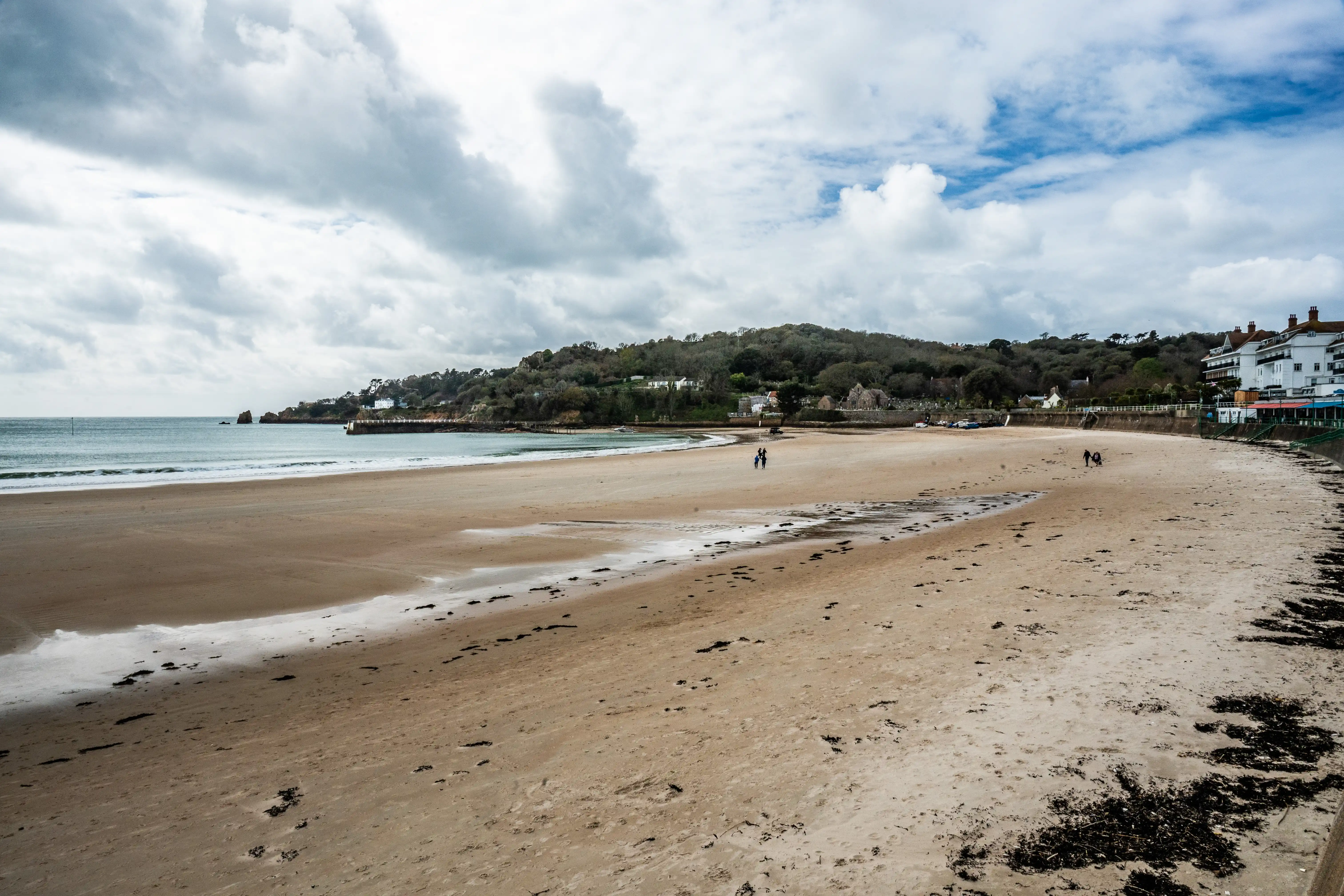 A wide, sandy beach with the sea on the left and buildings and a tree-covered hill in the background under a cloudy sky. Two people are walking on the sand.