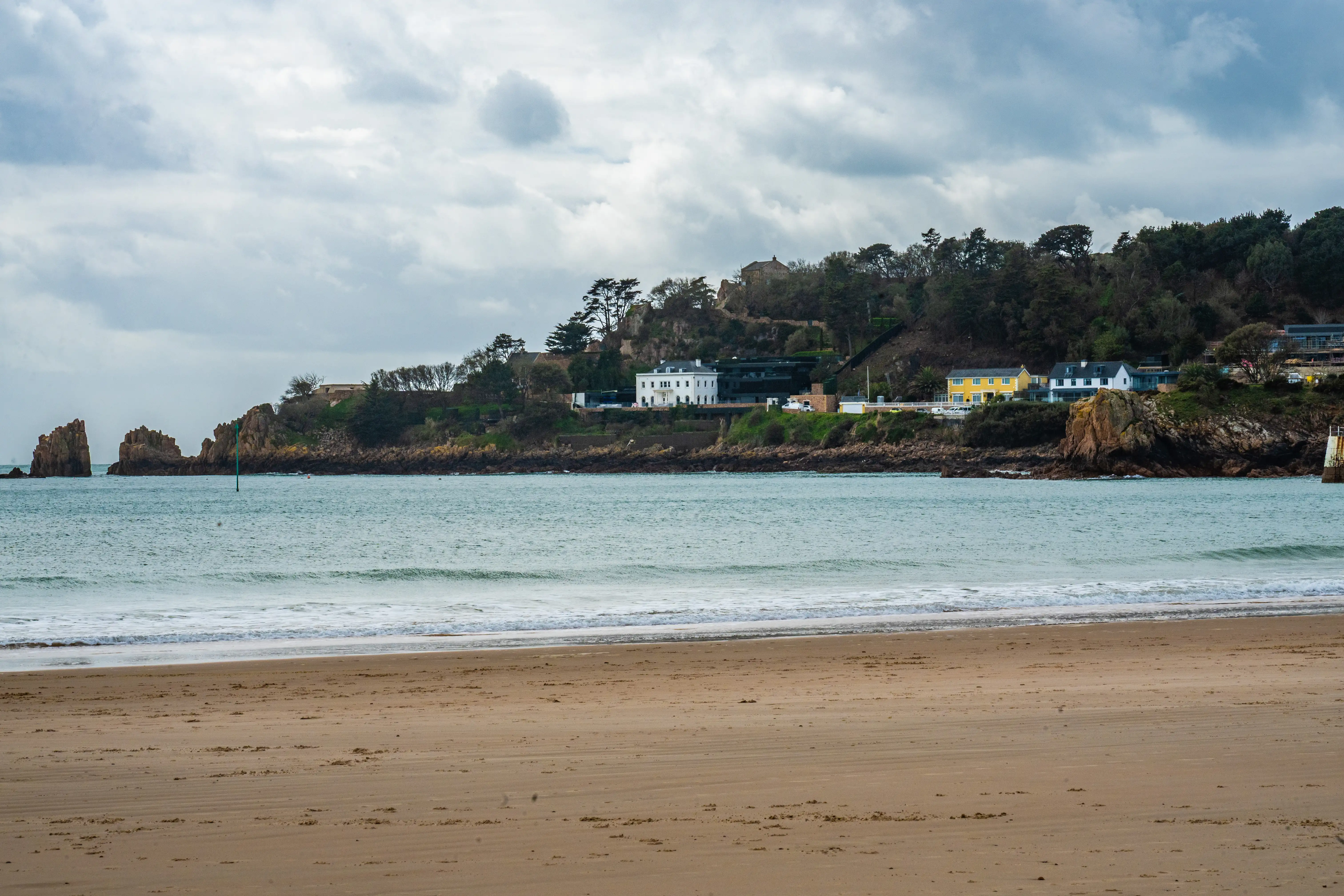 View of a beach with waves, overlooking a coastal hillside with several houses, including a large white villa, under a cloudy sky.