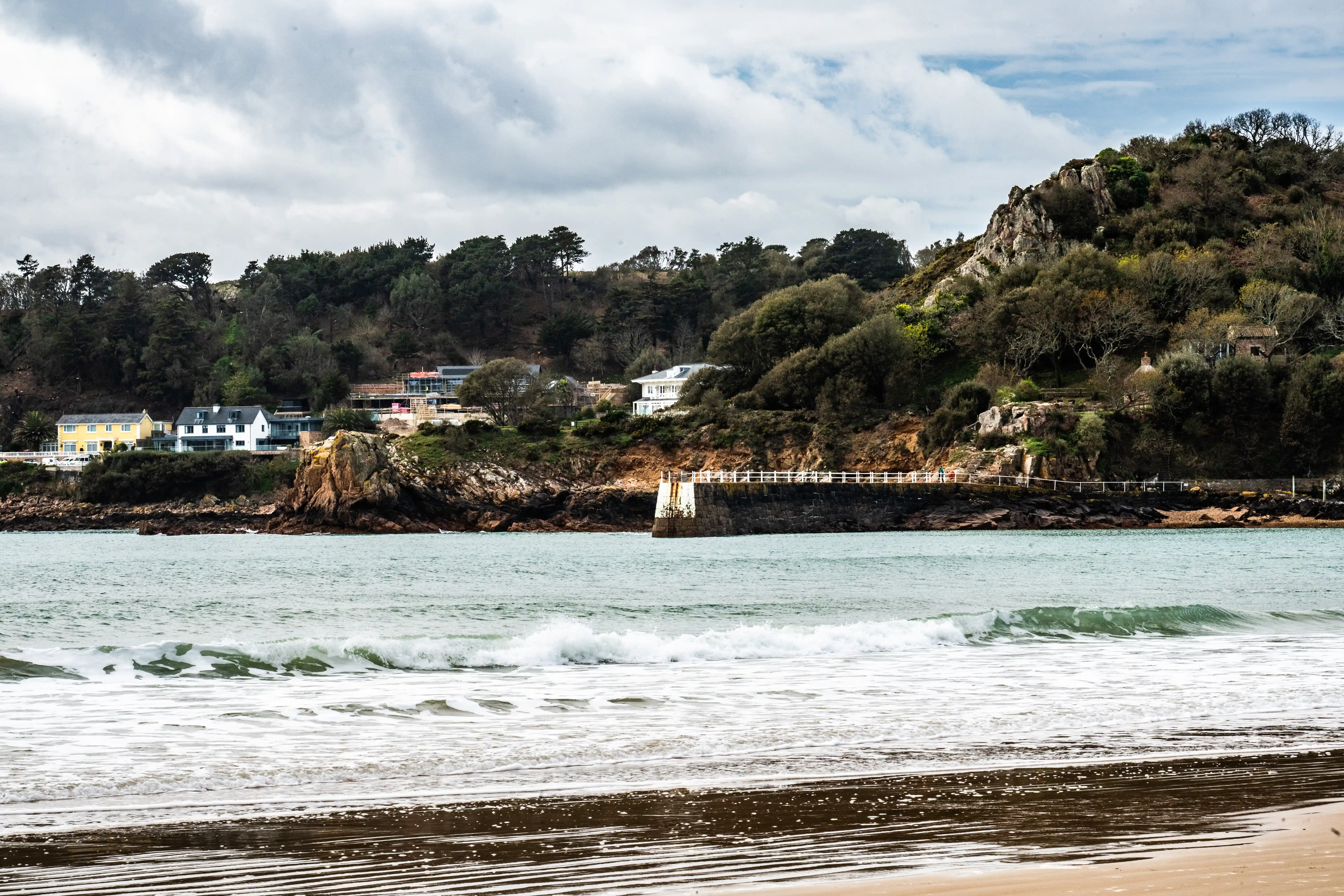 A wide shot of a coastline with several houses visible on a densely wooded hill. The sea in the foreground has small waves and white foam.