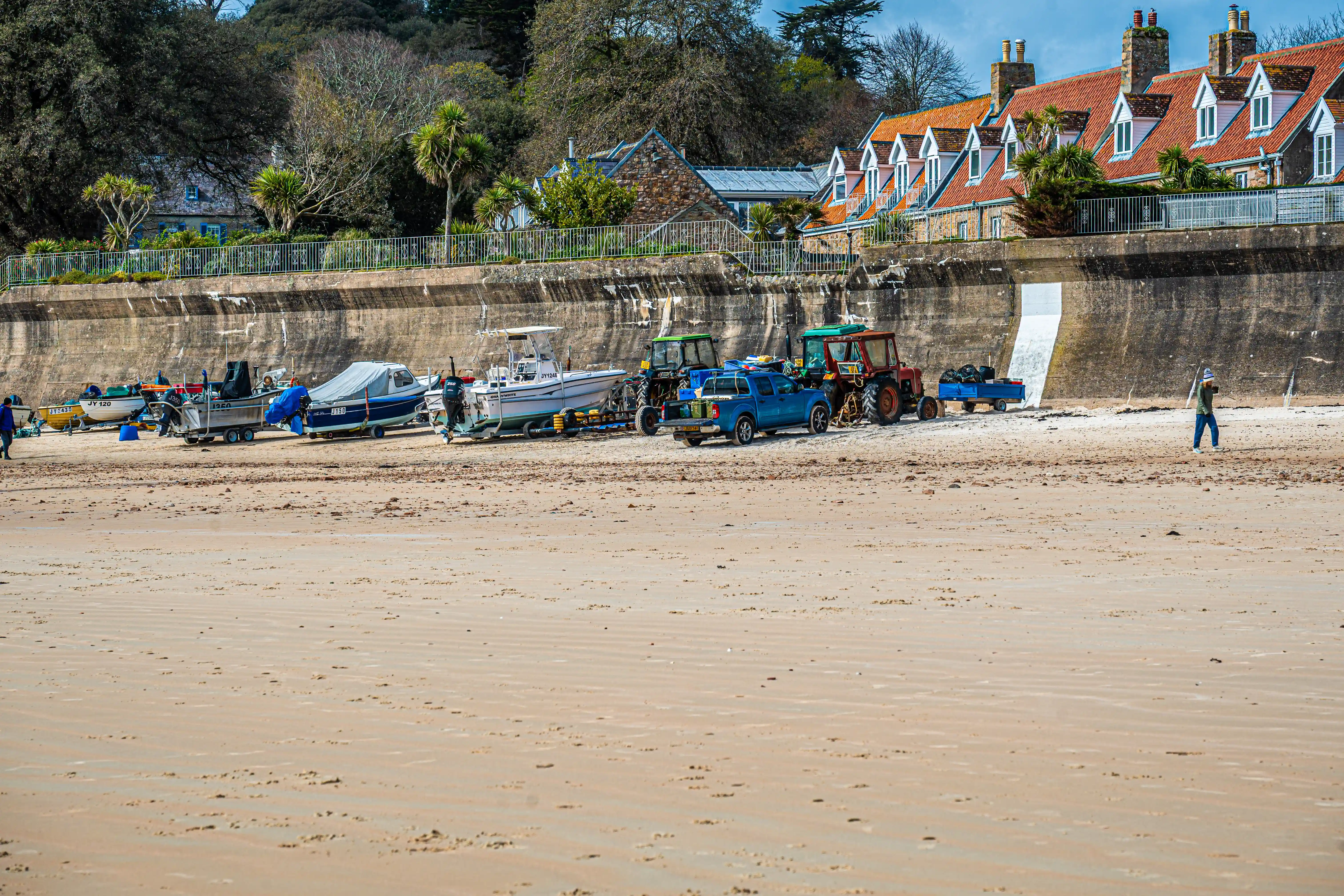 A sandy beach at low tide, with fishing boats on trailers, a blue pickup truck, and tractors. Behind a sea wall, a row of charming coastal houses with red roofs is visible, surrounded by green trees under a blue sky.