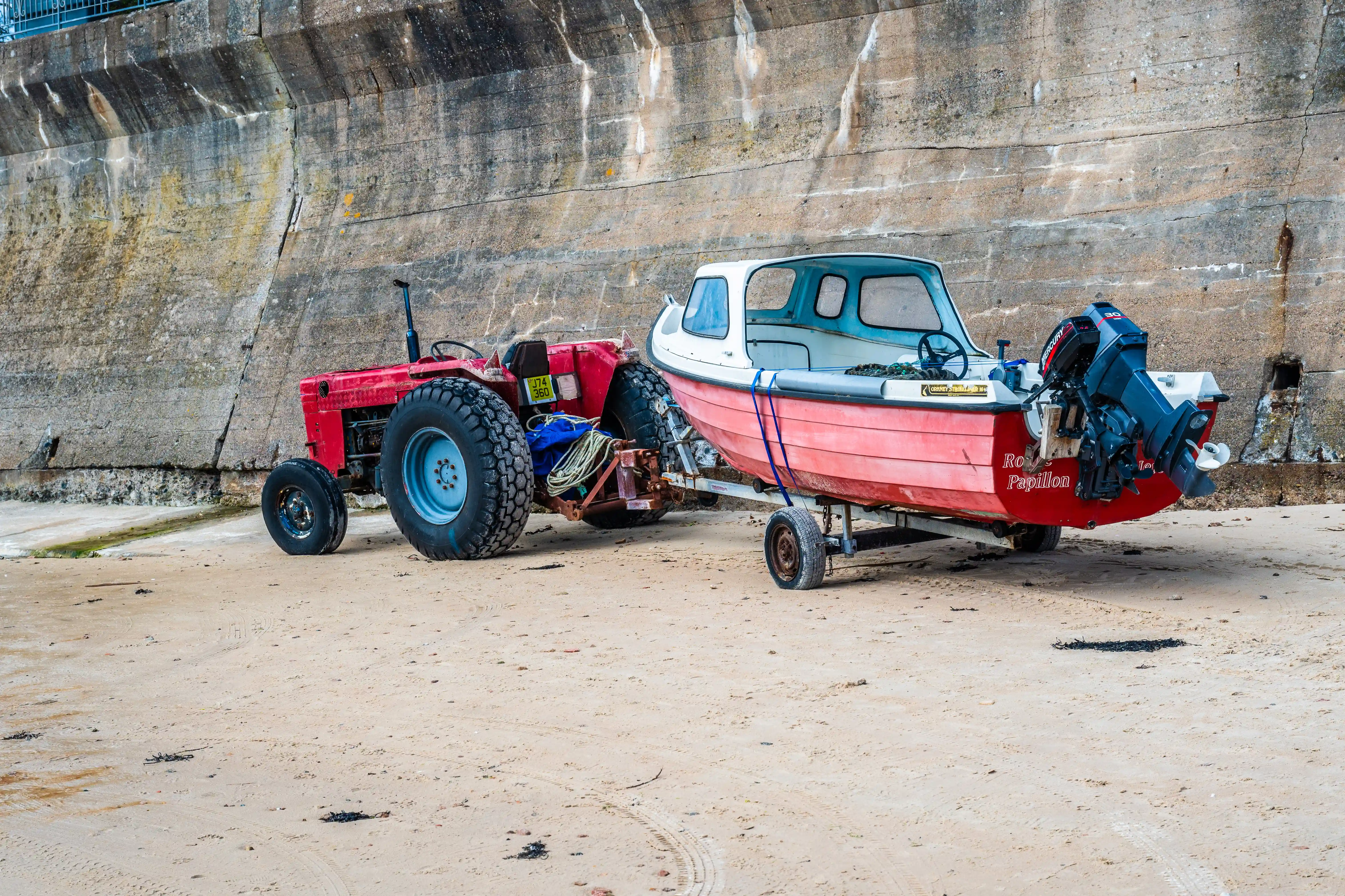 A red tractor is attached to a trailer with a red and white boat on it, positioned on a sandy beach beside a concrete wall.