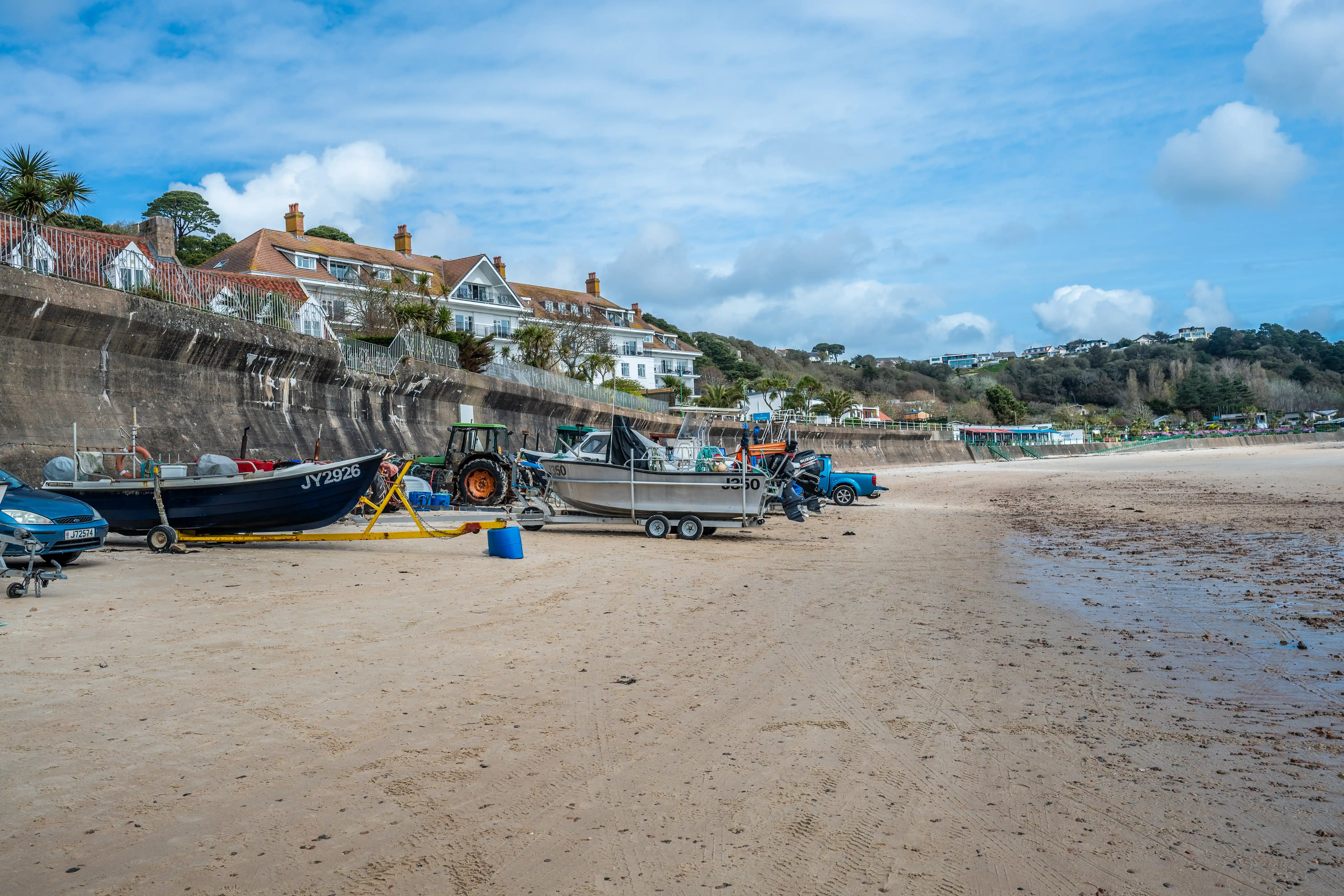 A wide shot of a sandy beach with various boats on trailers, a tractor, and a blue pickup truck. In the background, a concrete seawall protects a line of white buildings with red roofs, backed by a lush green hillside under a blue sky with scattered clouds.