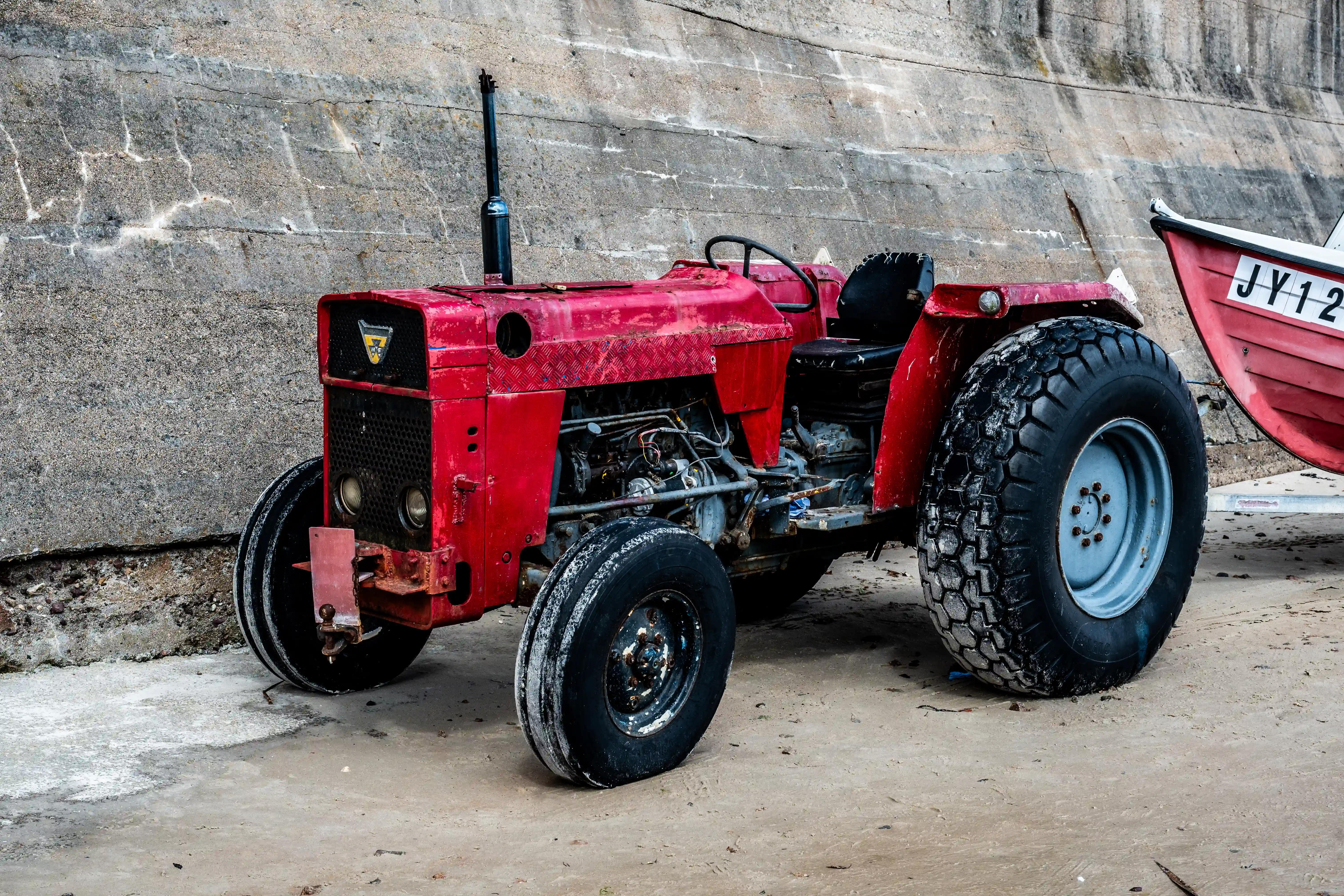 A red tractor parked on sand next to a concrete wall, with a red boat to its right.