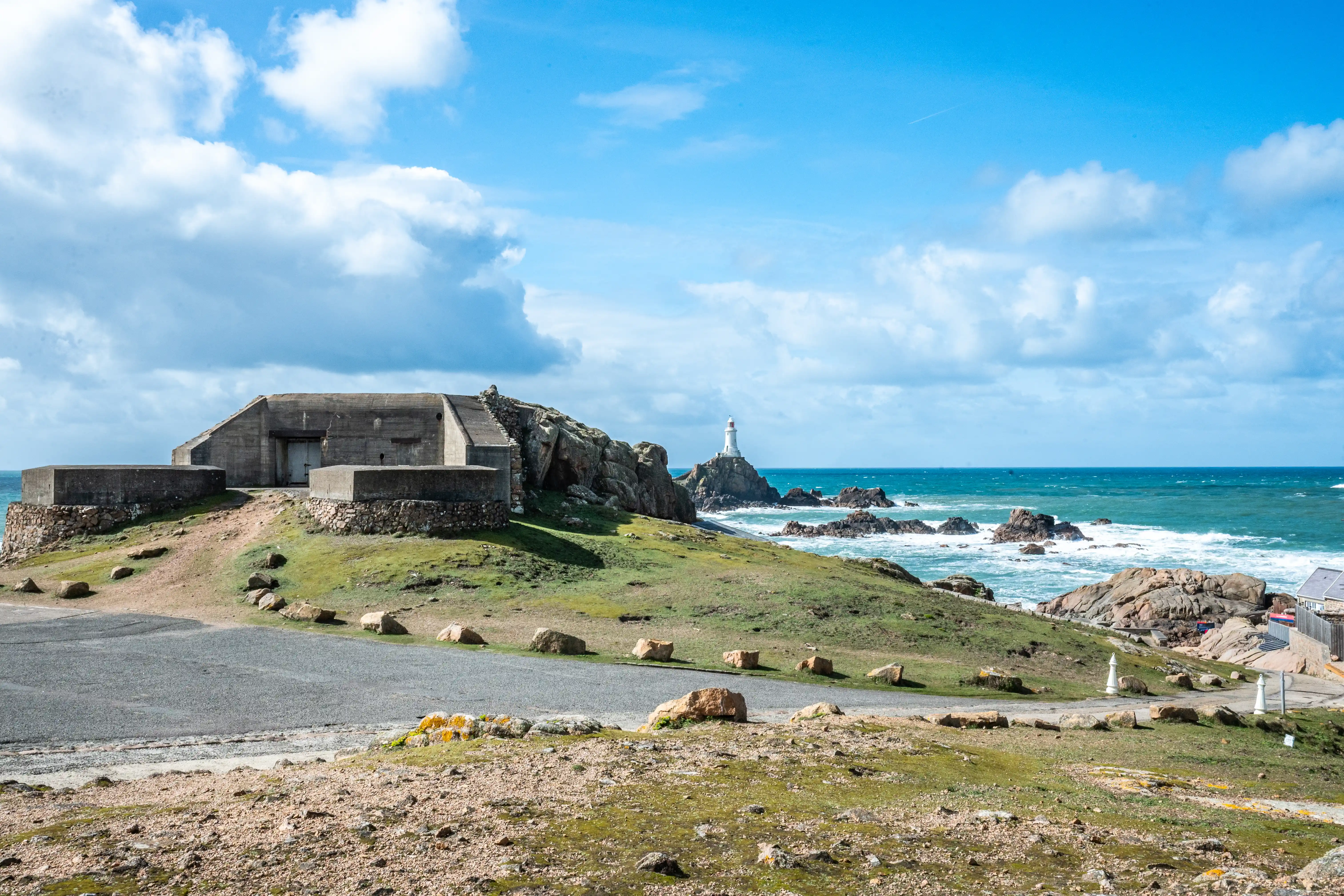 A panoramic view of a concrete military bunker on a grassy hill overlooking a turbulent sea. A white lighthouse is visible on a distant rocky island.