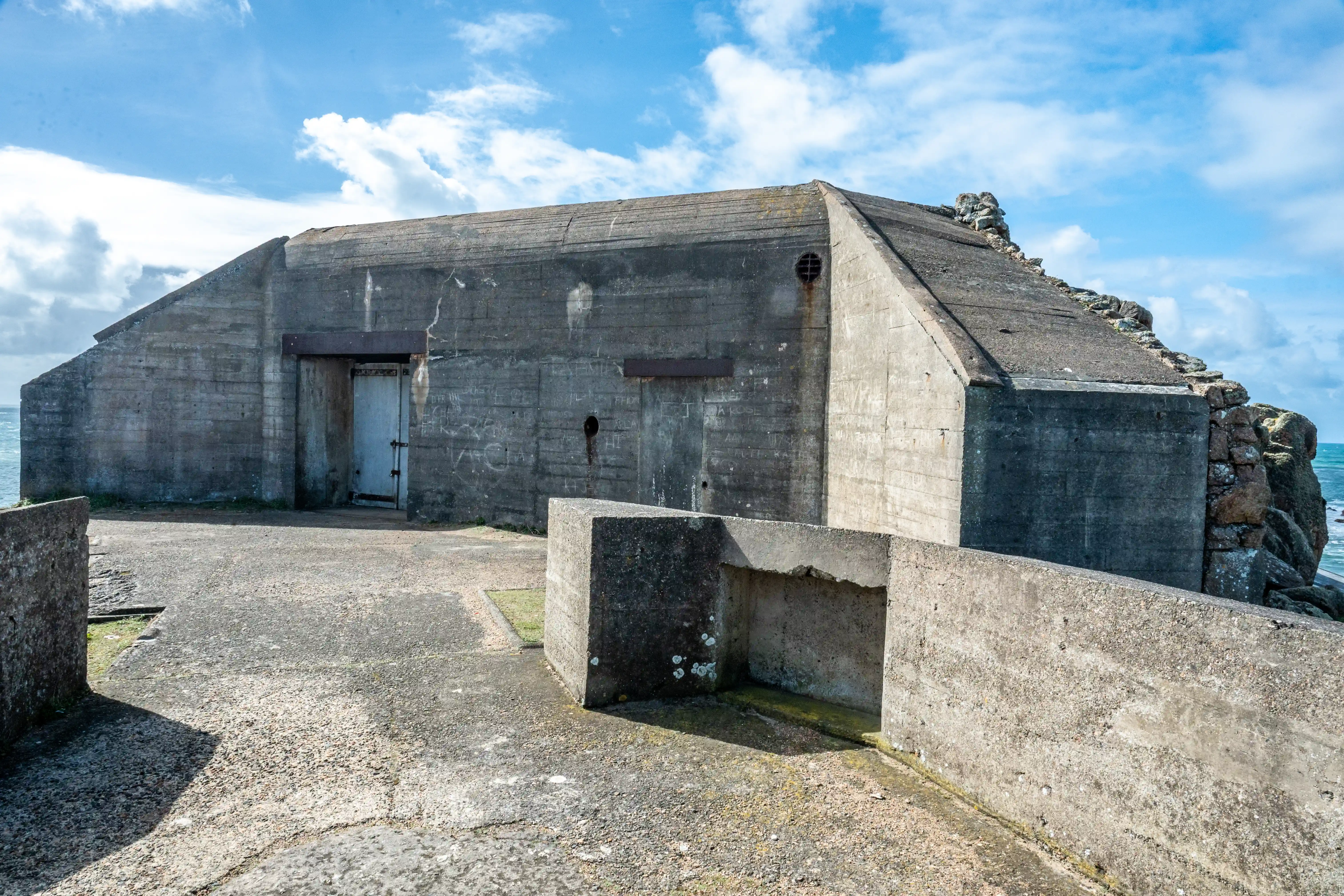A view of a substantial, aged concrete military bunker with a metal door, located beside a rugged coastline under a dynamic sky with white clouds.