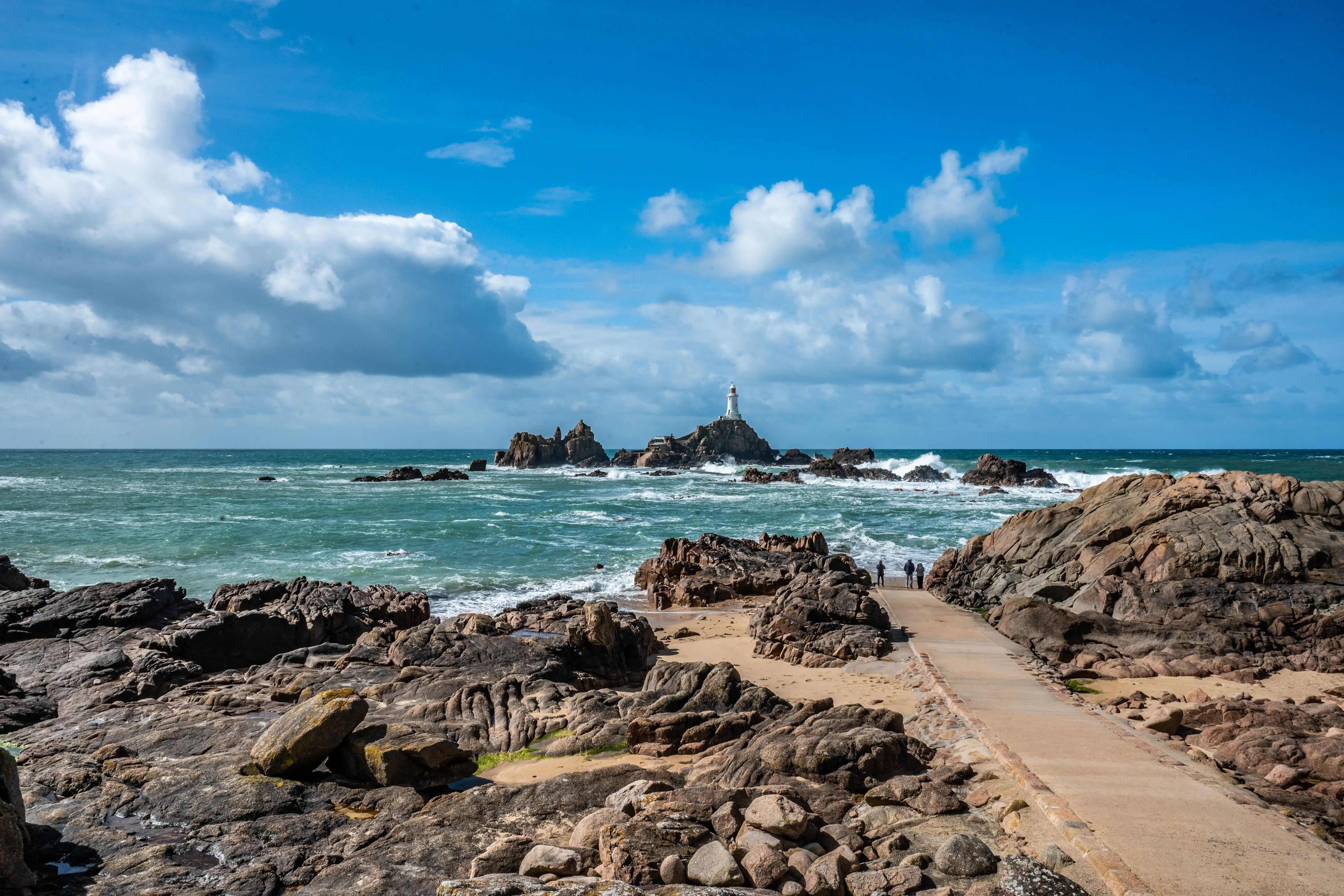 A lighthouse stands on a rocky outcrop in a choppy sea, with a concrete path leading towards it from a rocky foreground under a blue sky with white clouds.