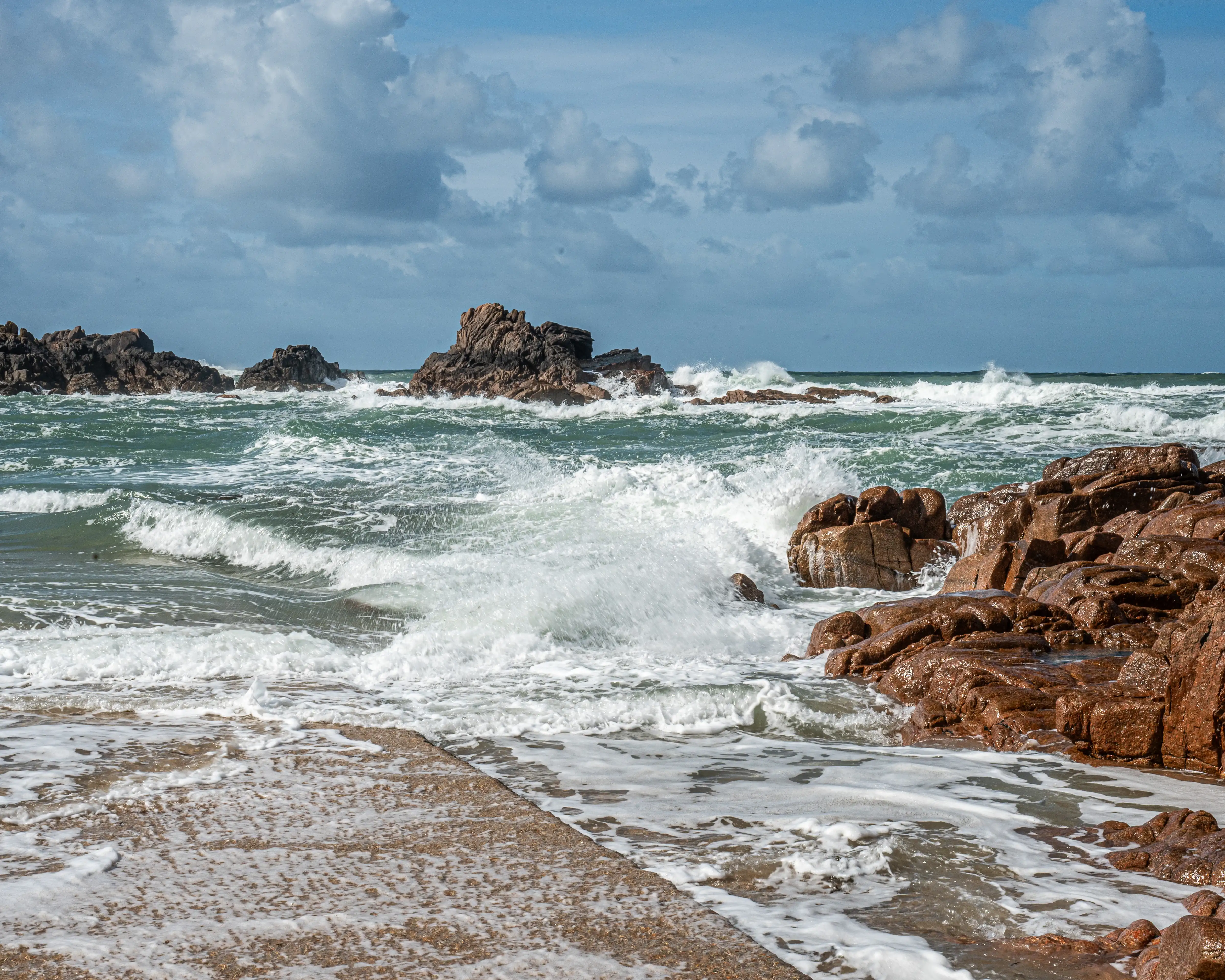  A dramatic seascape captures the power of the ocean as waves crash against a rugged, rocky coastline under a partially cloudy sky. The foreground features a concrete pathway leading into the sea, with foam and water lapping at its edges. The sea is a vibrant green-blue, churned into white foam by the powerful swells. Dark, imposing rock formations jut out of the water in the mid-ground, while in the distance, the horizon is marked by further rocky outcrops and a hazy sea. The sky is a mix of blue with fluffy white and grey clouds, suggesting a dynamic and possibly windy day.