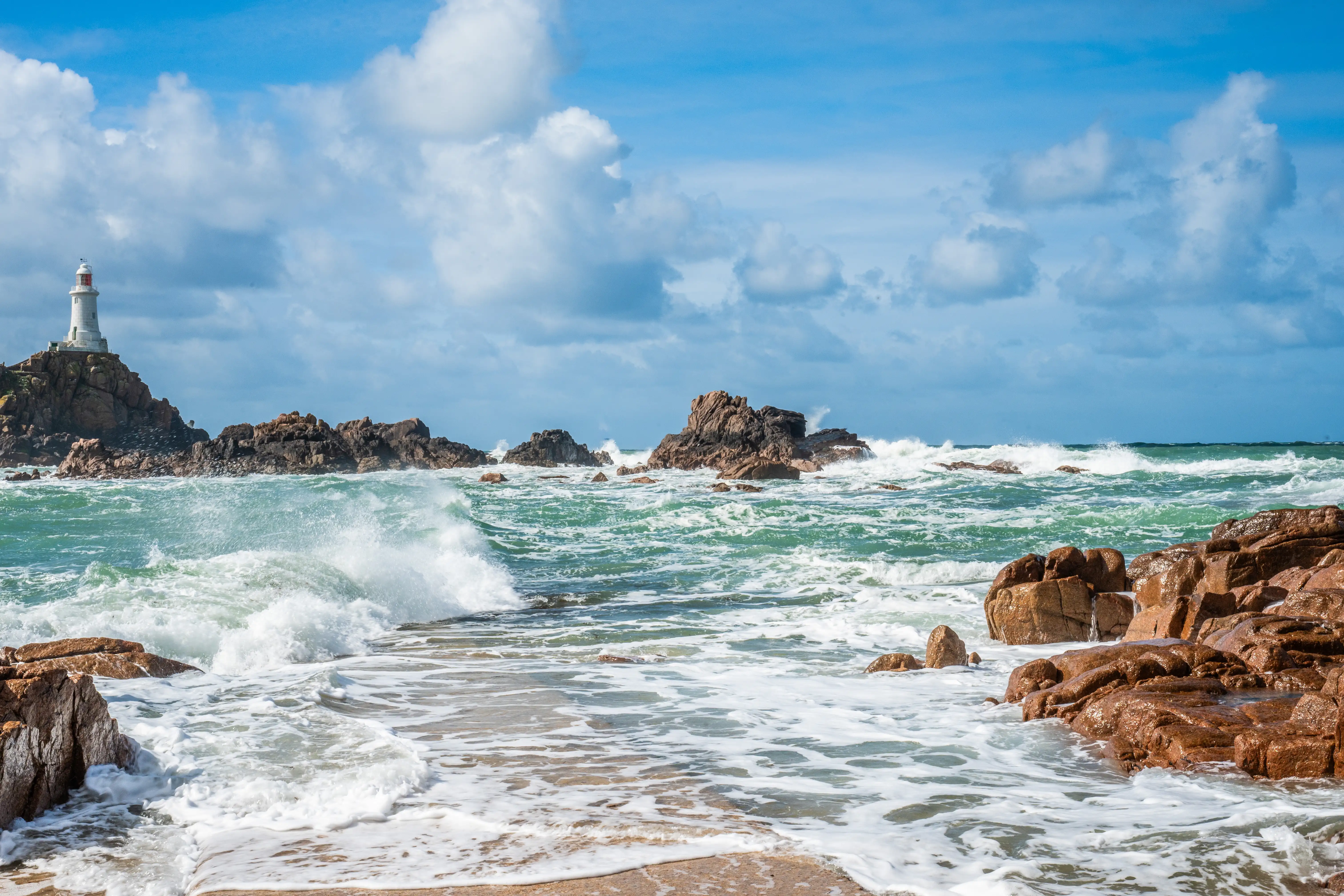  A white lighthouse stands tall on a rocky island, with rough, turquoise waves crashing against the shore and surrounding red-brown rocks. The sky is a bright blue with fluffy white clouds.