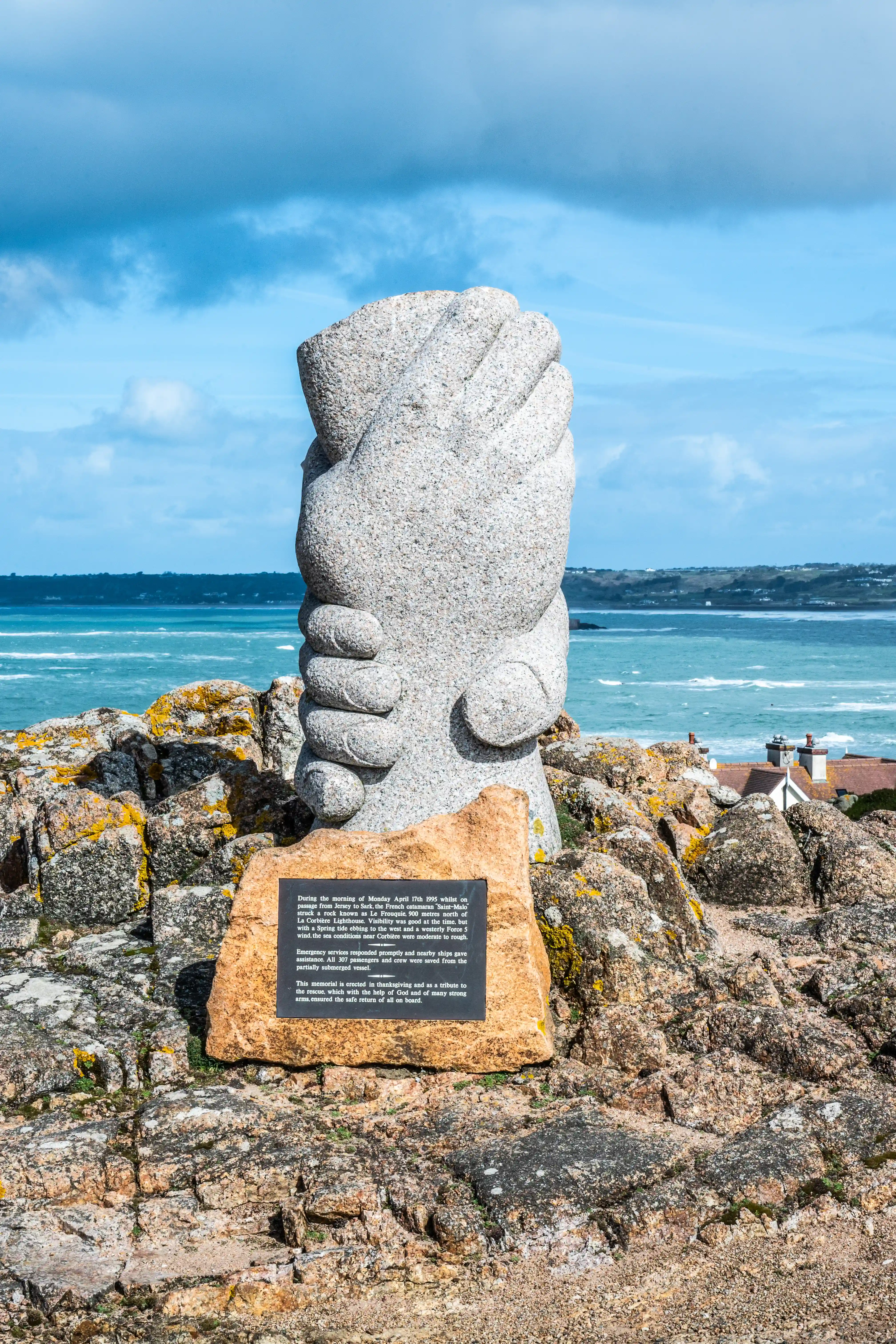 A grey granite sculpture of two hands clasped together is positioned on a rugged, rocky coastline. In the background, the turquoise sea stretches to the horizon under a partly cloudy blue sky. A small plaque at the base of the sculpture provides details of a maritime rescue.
