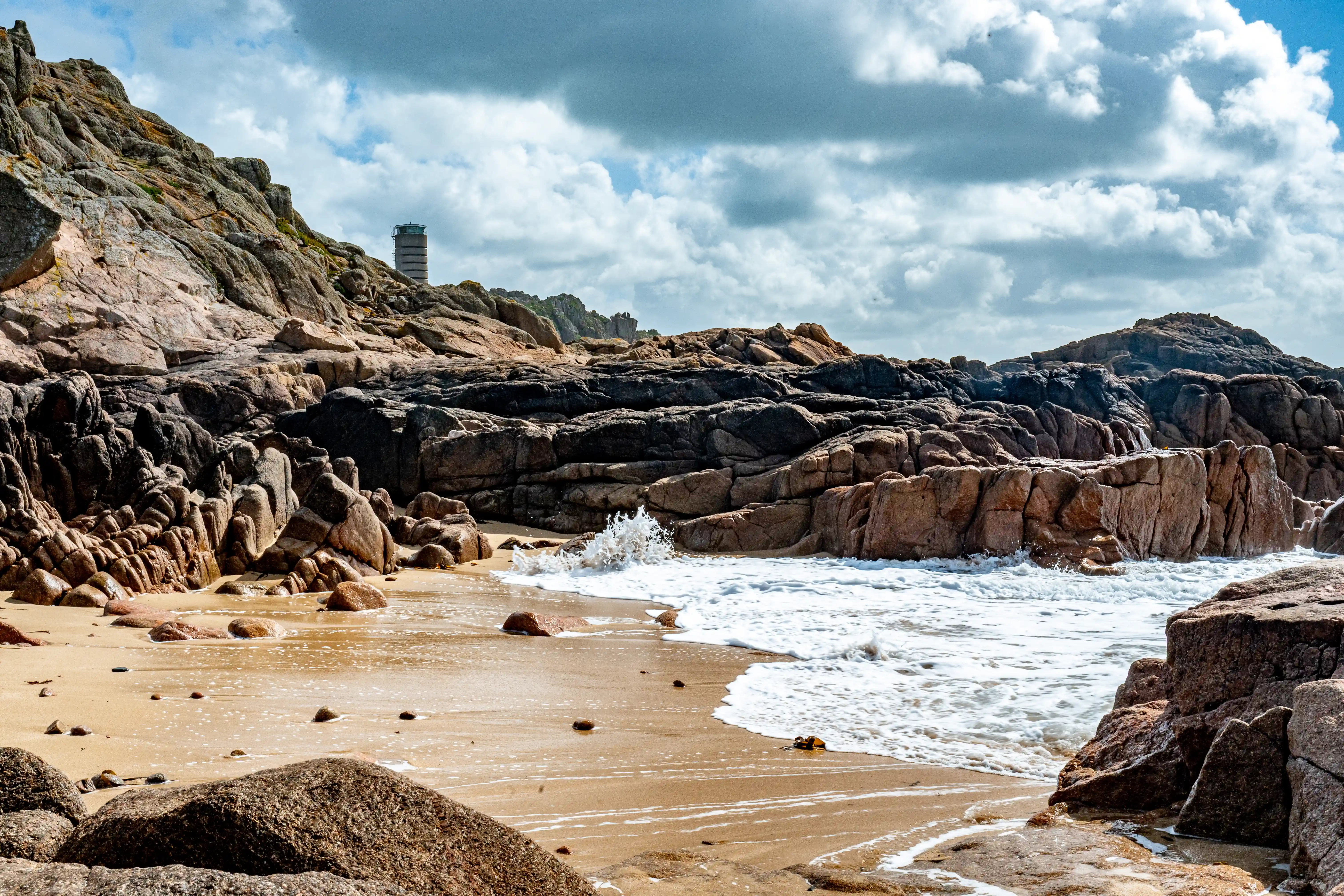  A natural view of a rocky coastline with a beach and the sea. A modern cylindrical tower structure, likely a lighthouse, is visible on a distant hill under a partly cloudy sky. Waves are crashing onto the shore, creating white foam.