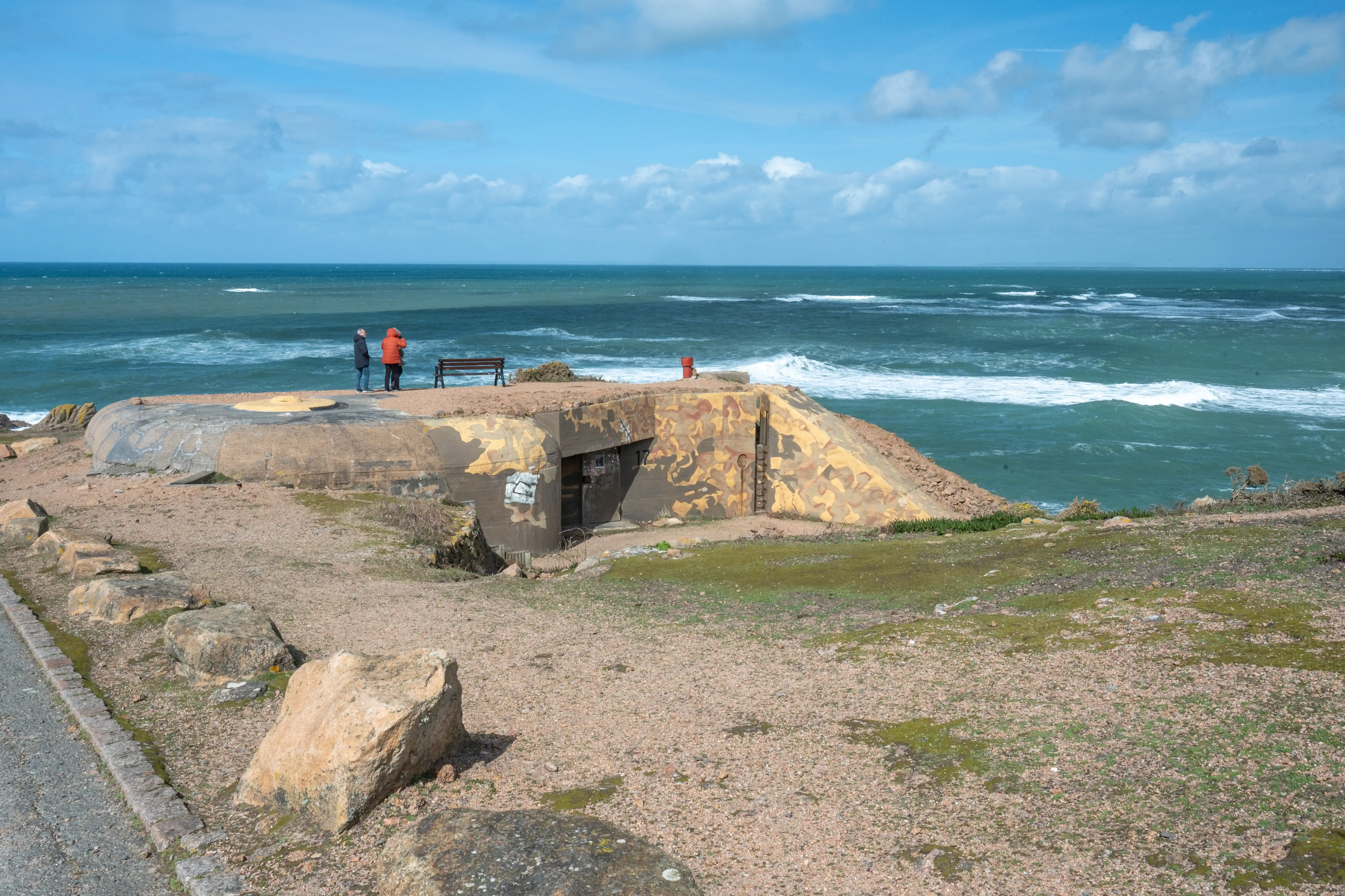 A camouflaged WWII bunker on a cliff overlooking a rough sea, with two people standing nearby and a wooden bench.