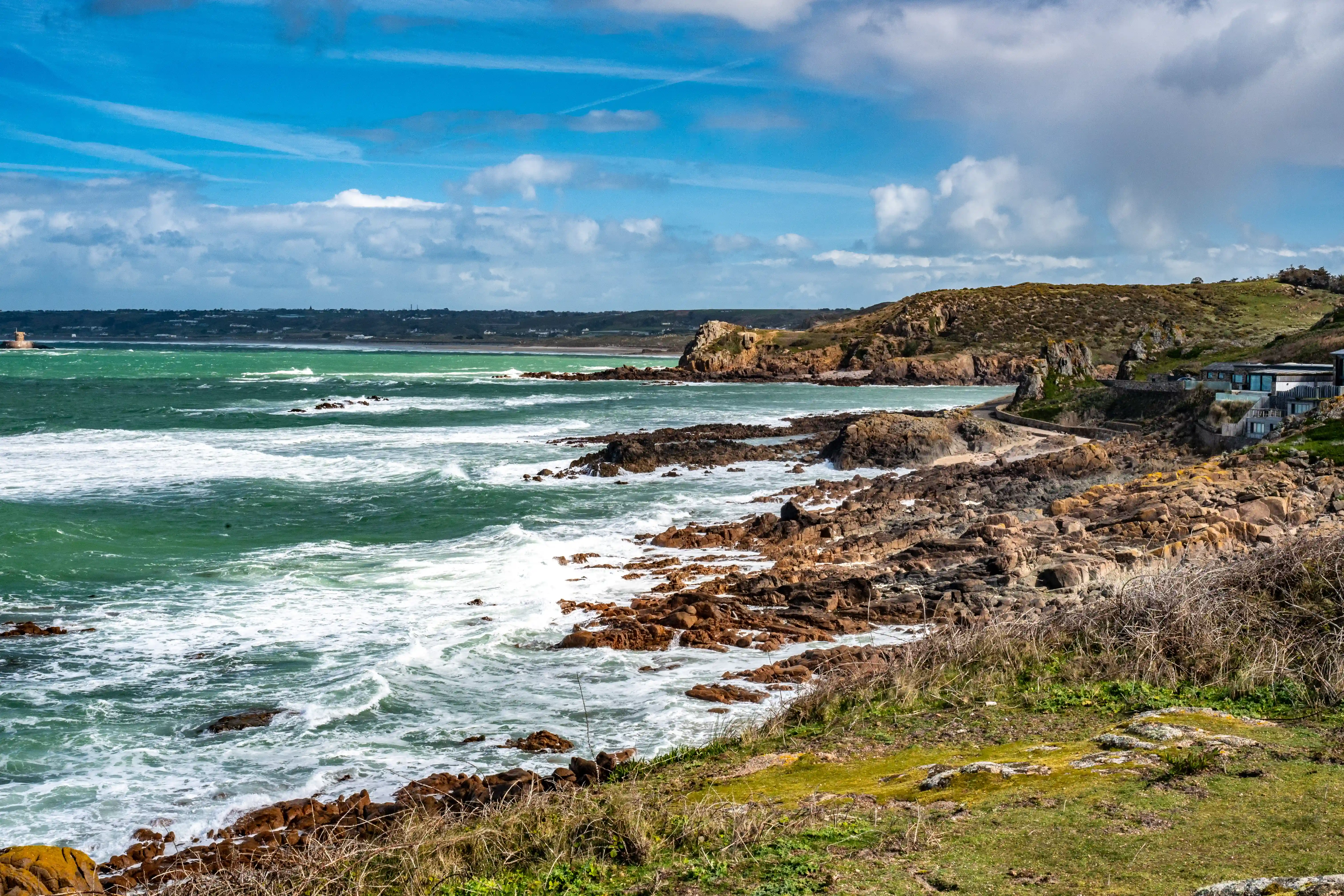 A dramatic coastal landscape in Point Corbiere, Jersey, showing rough seas, rocky shores, a distant town, and modern homes overlooking the ocean under a partly cloudy blue sky.