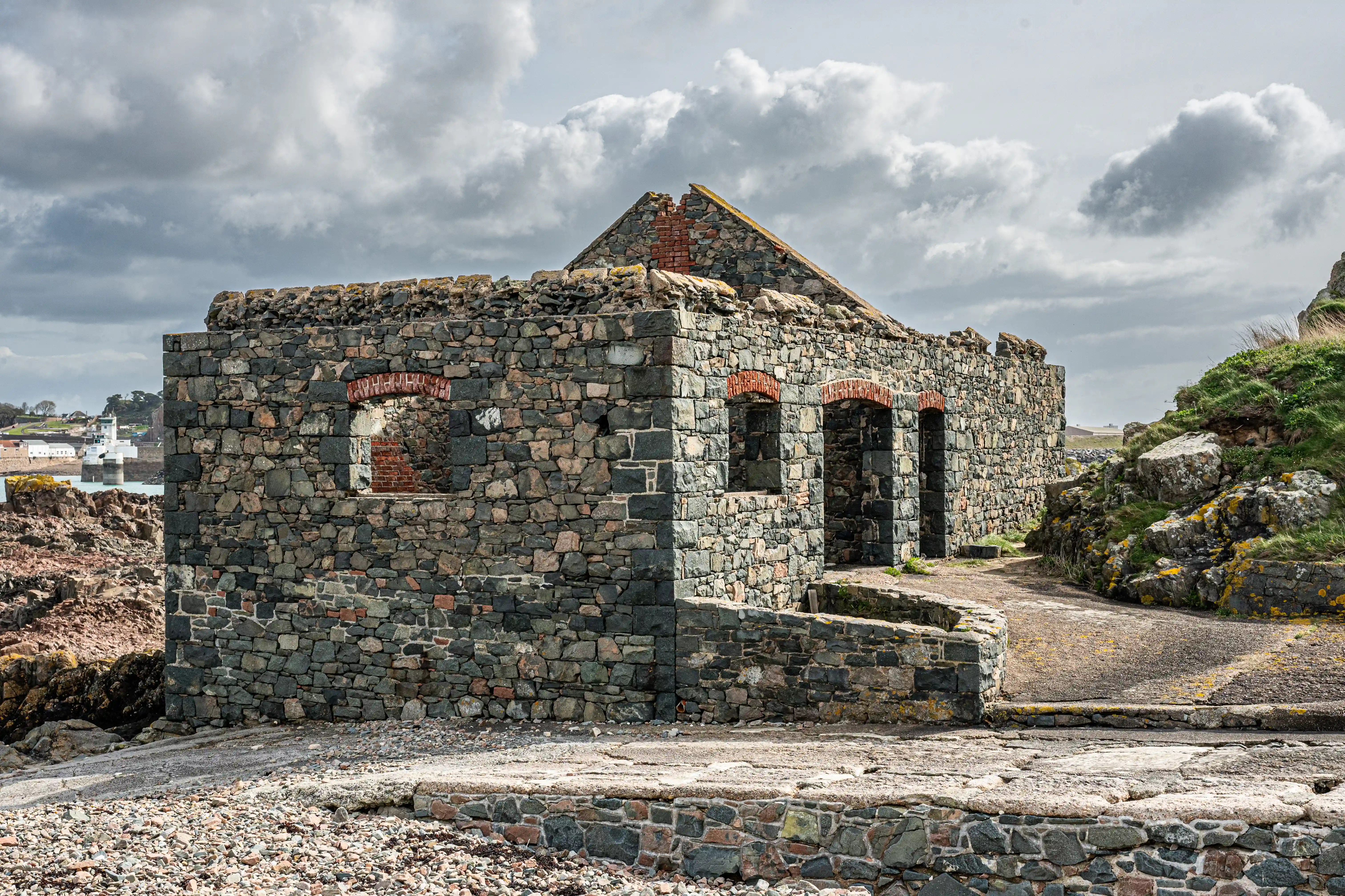 A weathered, stone-built ruin with arched window openings and a damaged roof, situated on a stony beach with a cloudy sky overhead.