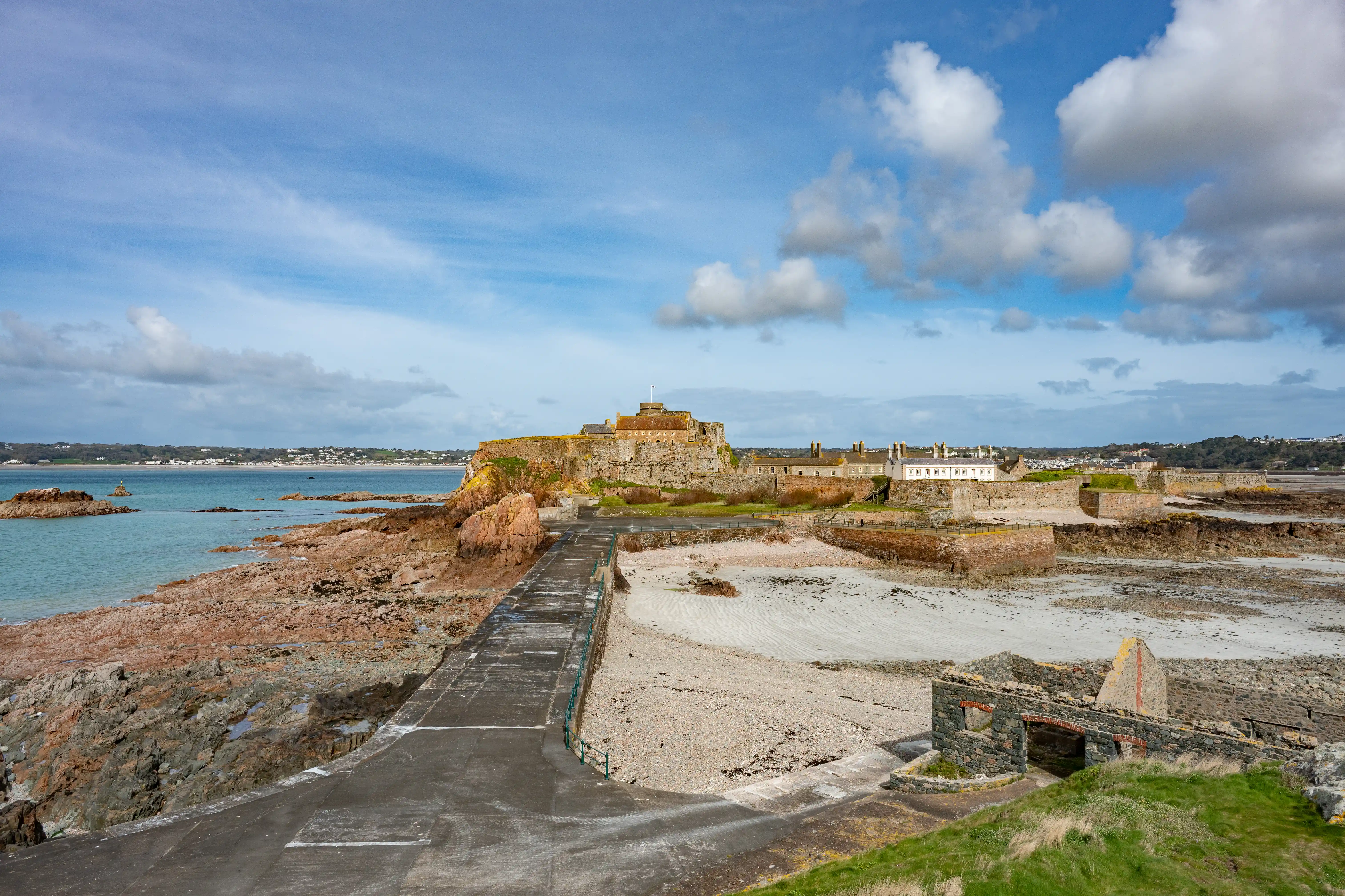 A panoramic view of a stone fortress and harbour on a rocky coastline, with a concrete path leading to it. The sea is to the left, and a town is visible in the distance under a cloudy blue sky.