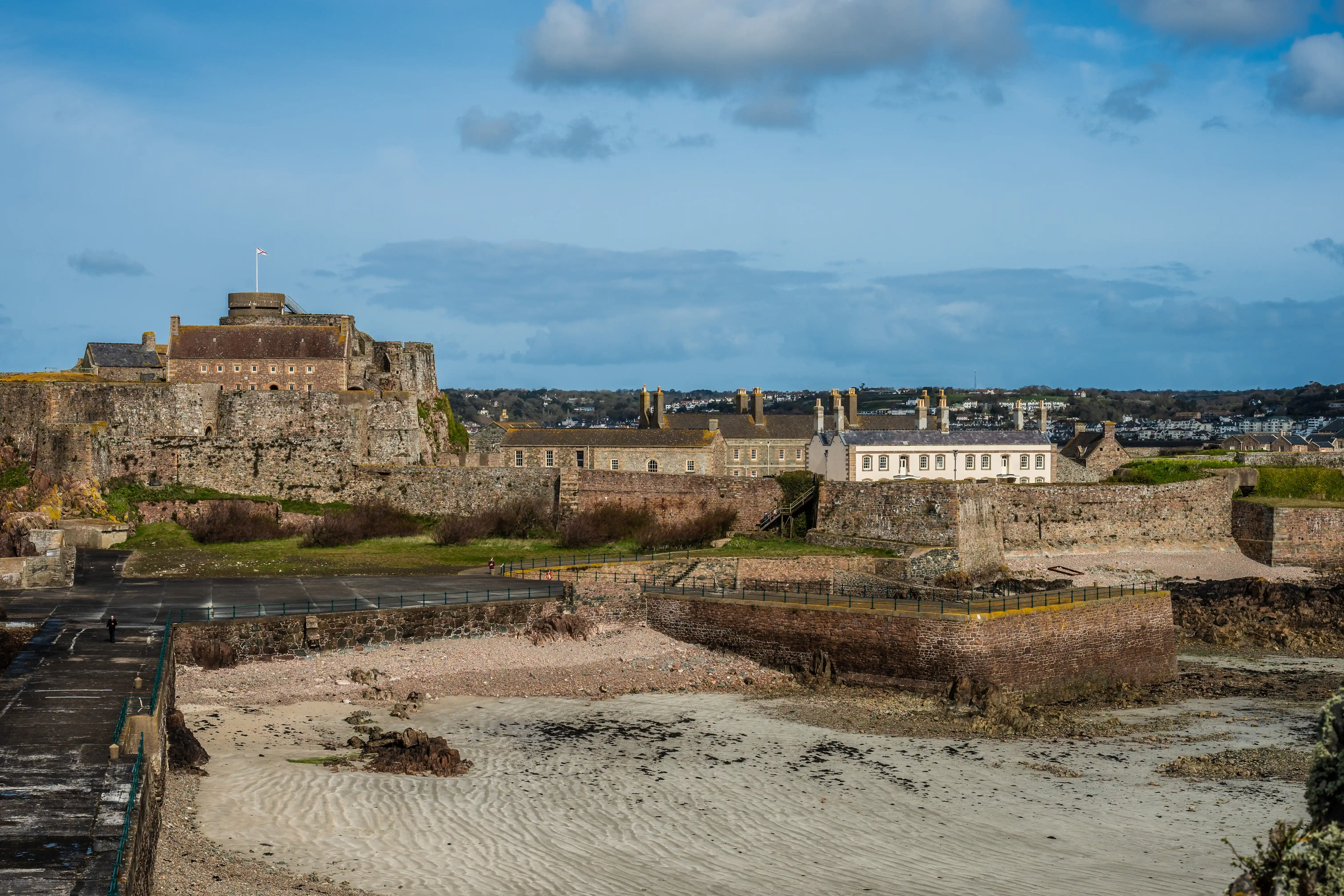 Wide shot of Elizabeth Castle, Jersey, with its stone walls and battlements, situated on a rocky outcrop overlooking a sandy beach. Buildings within the castle complex and a town in the background are visible under a sky with scattered clouds.