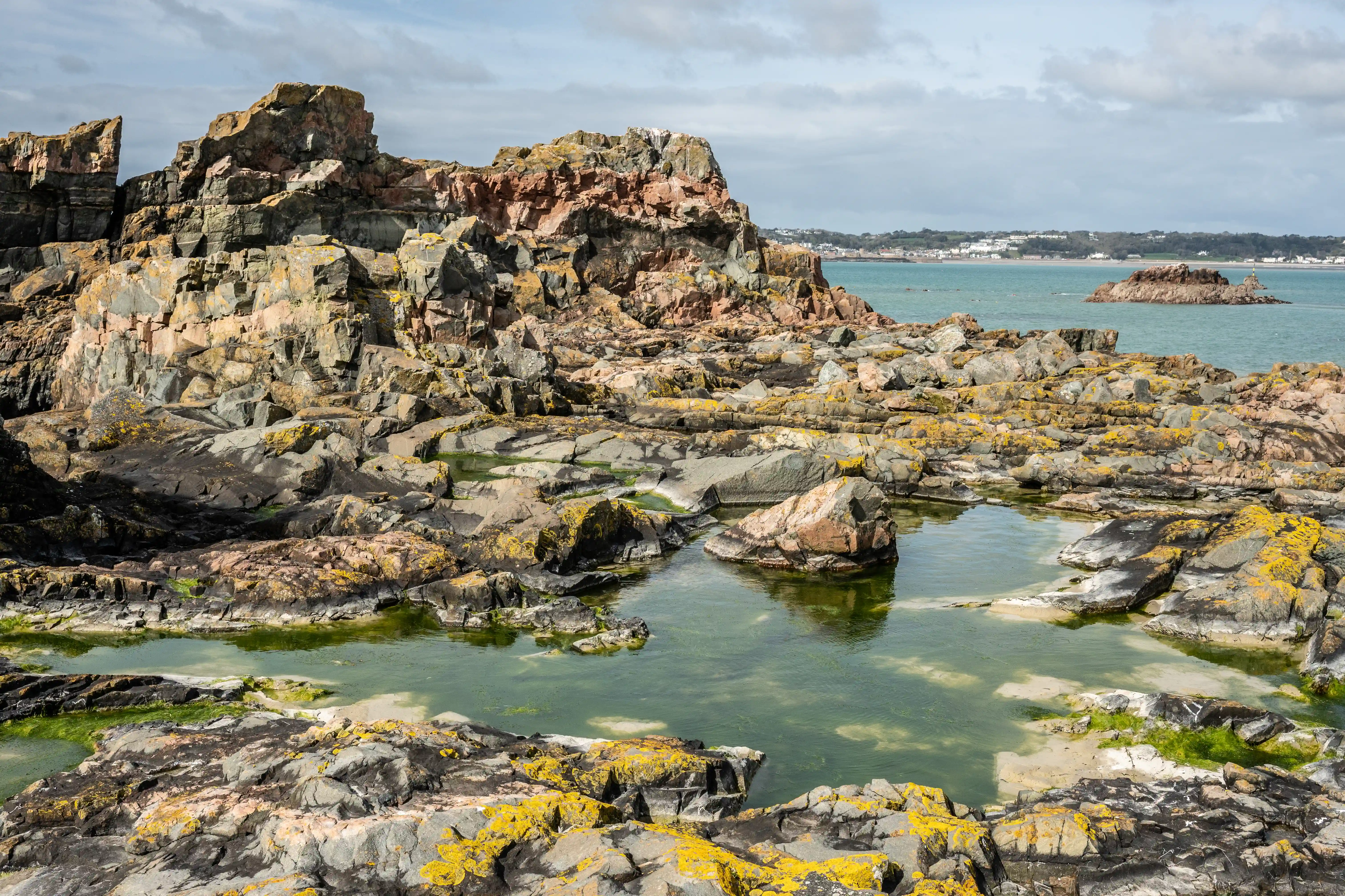 A panoramic view of a stone fortress and harbour on a rocky coastline, with a concrete path leading to it. The sea is to the left, and a town is visible in the distance under a cloudy blue sky.