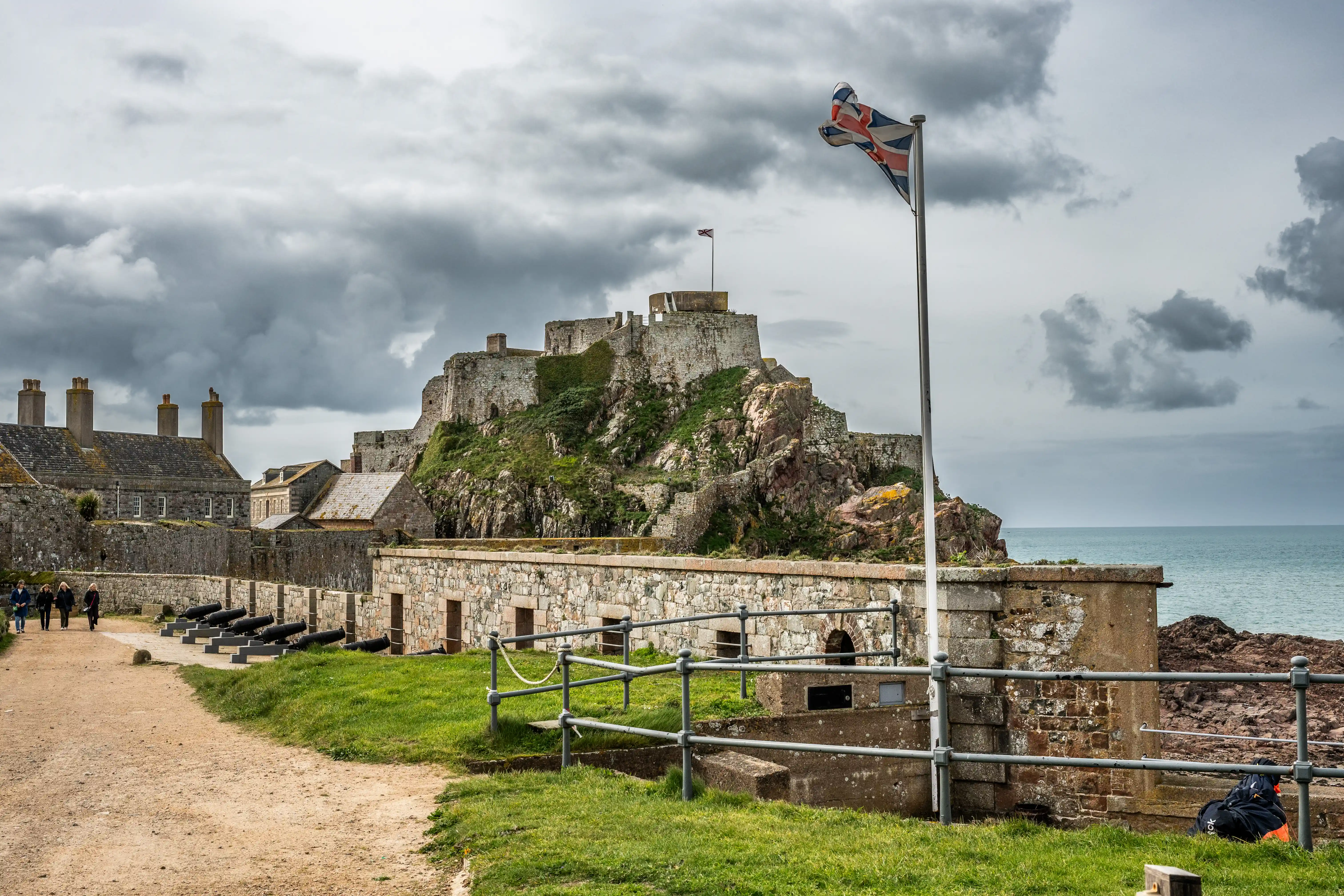 A historic stone castle on a rocky hill under a cloudy sky, with a Union Jack flag flying. Cannons are visible along a stone wall in the foreground, and the sea is in the distance.