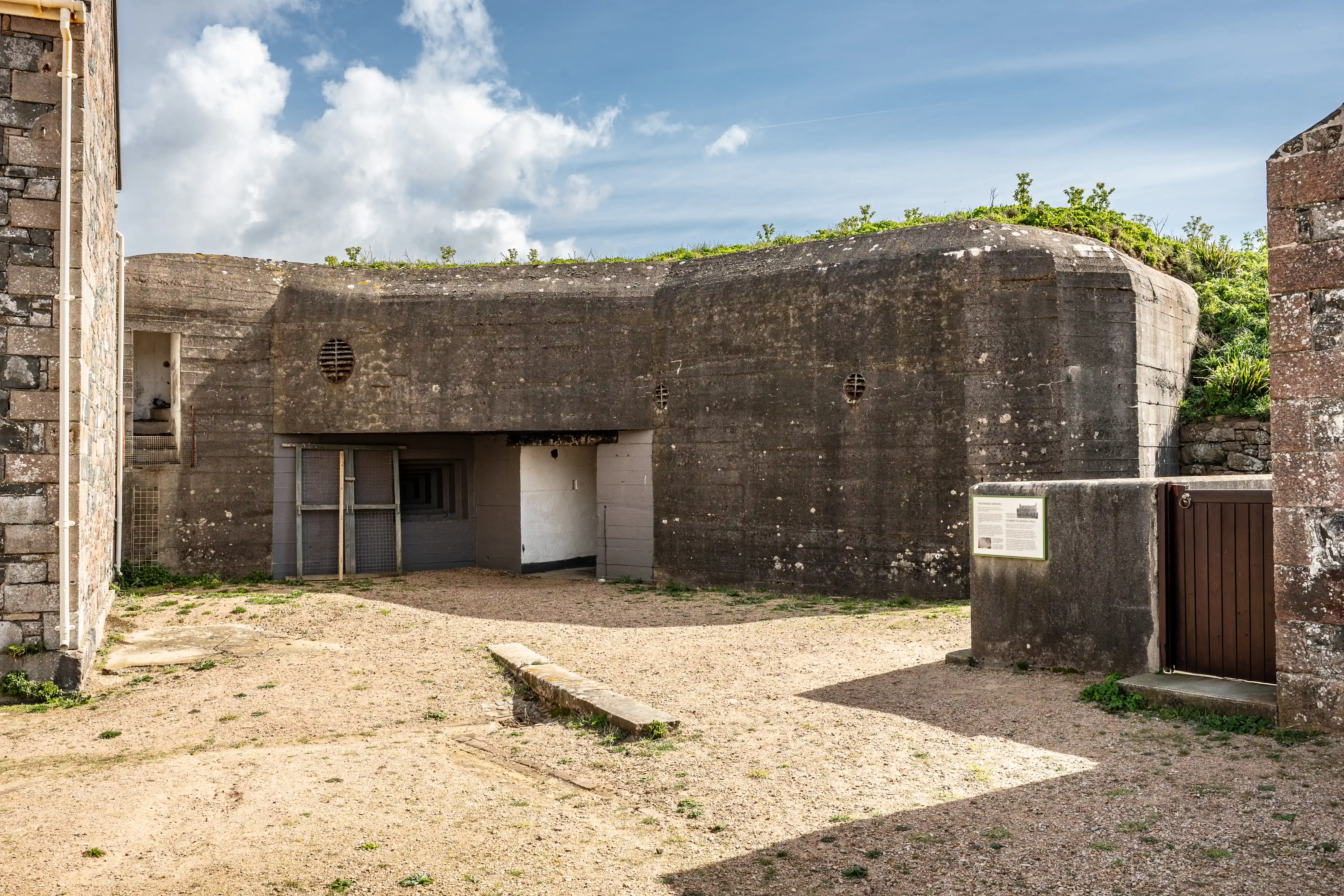 A concrete coastal defence bunker with multiple entrances and ventilation grilles, set in a gravelled area with stone buildings to the left and right, under a cloudy blue sky.