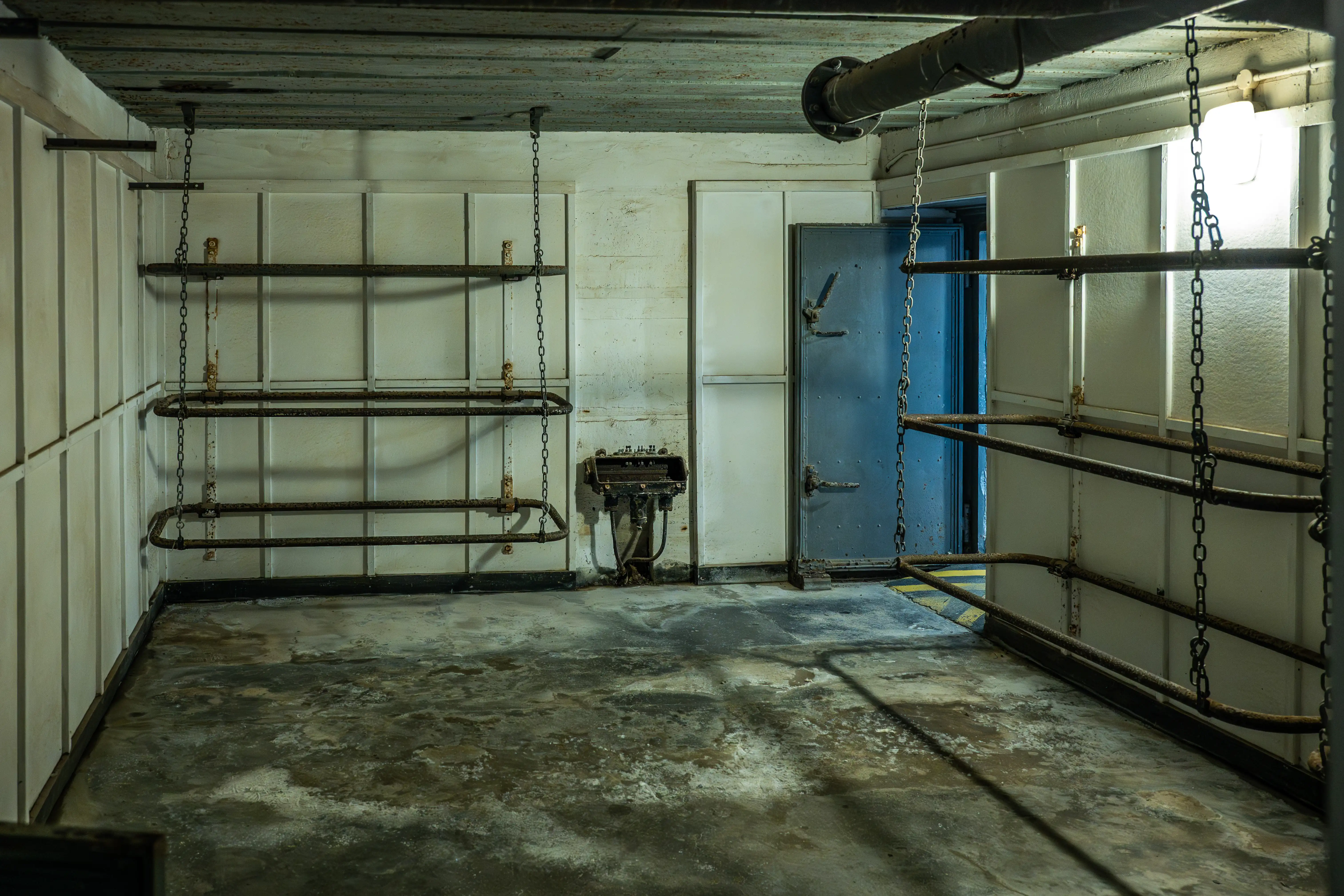 Interior view of a disused bunker room with concrete floor, white panelled walls, rusty metal shelves hanging from chains, and a heavy blue metal door.