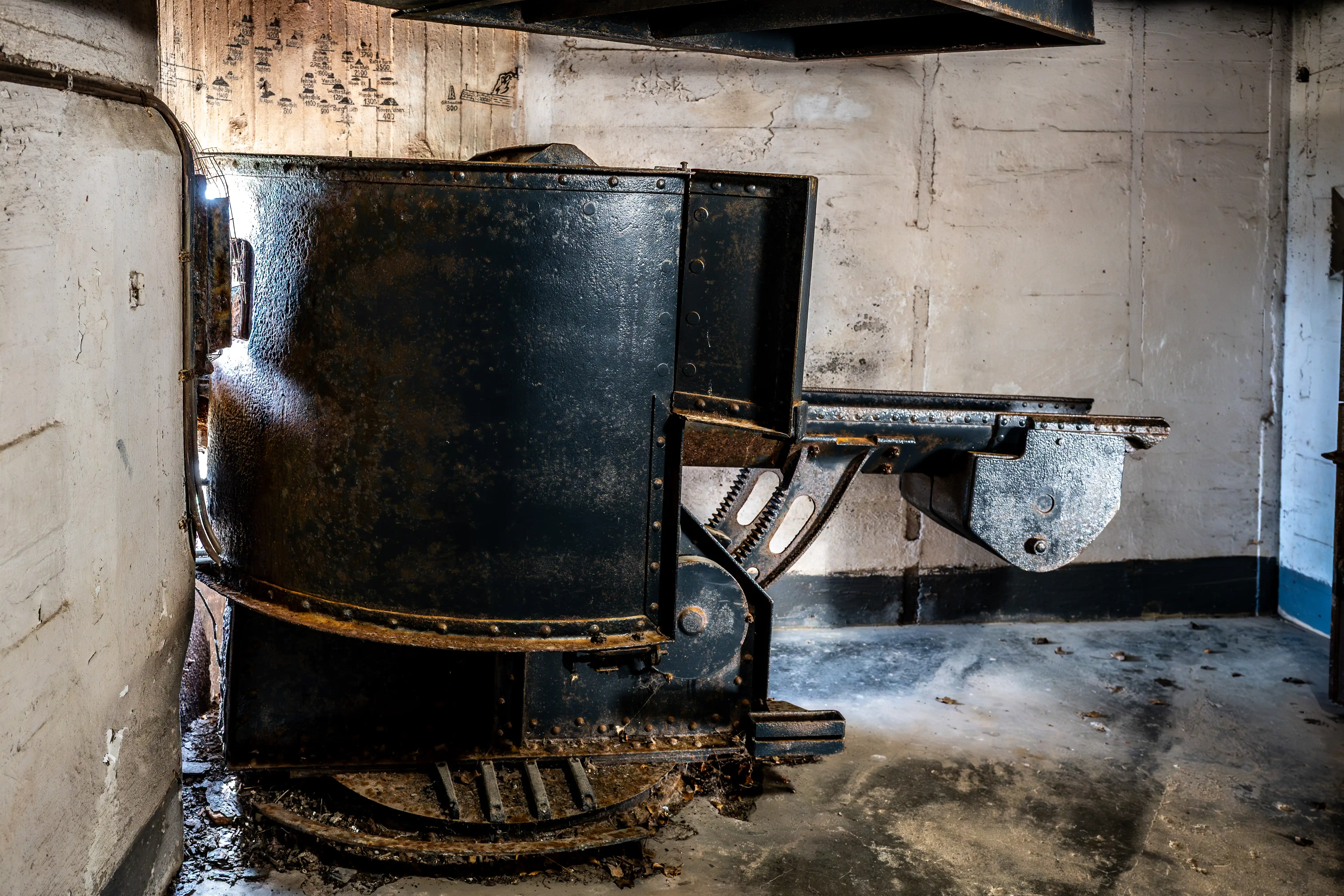 Close-up of a weathered, dark metal industrial mechanism inside a concrete bunker, with rust and faint markings on the walls.