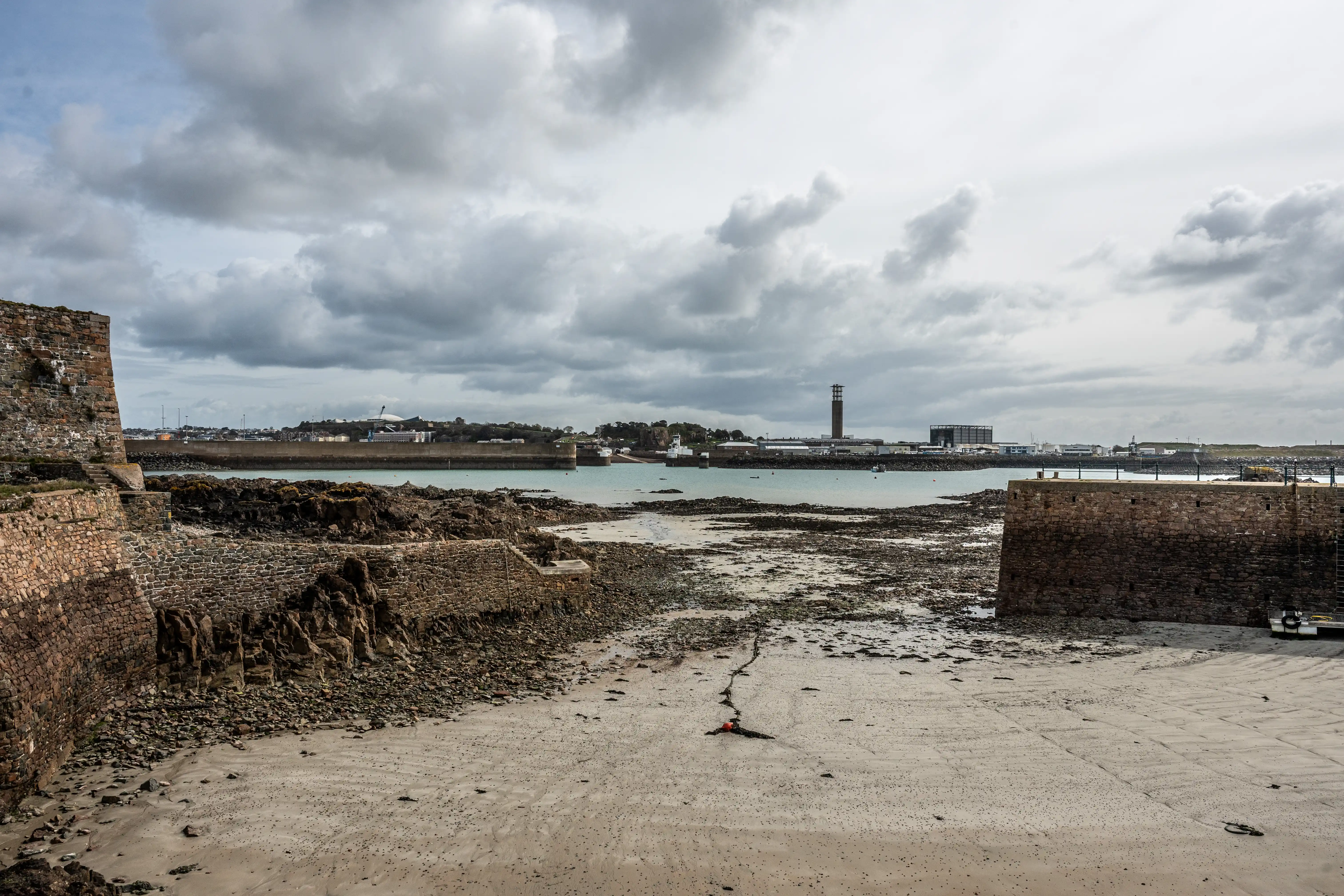 A wide-angle shot of a harbour with a stone wall and rocky embankment in the foreground, leading to a body of water. The background shows a town or industrial area with a tall, distinctive tower and several other structures, all under a sky filled with heavy grey clouds.