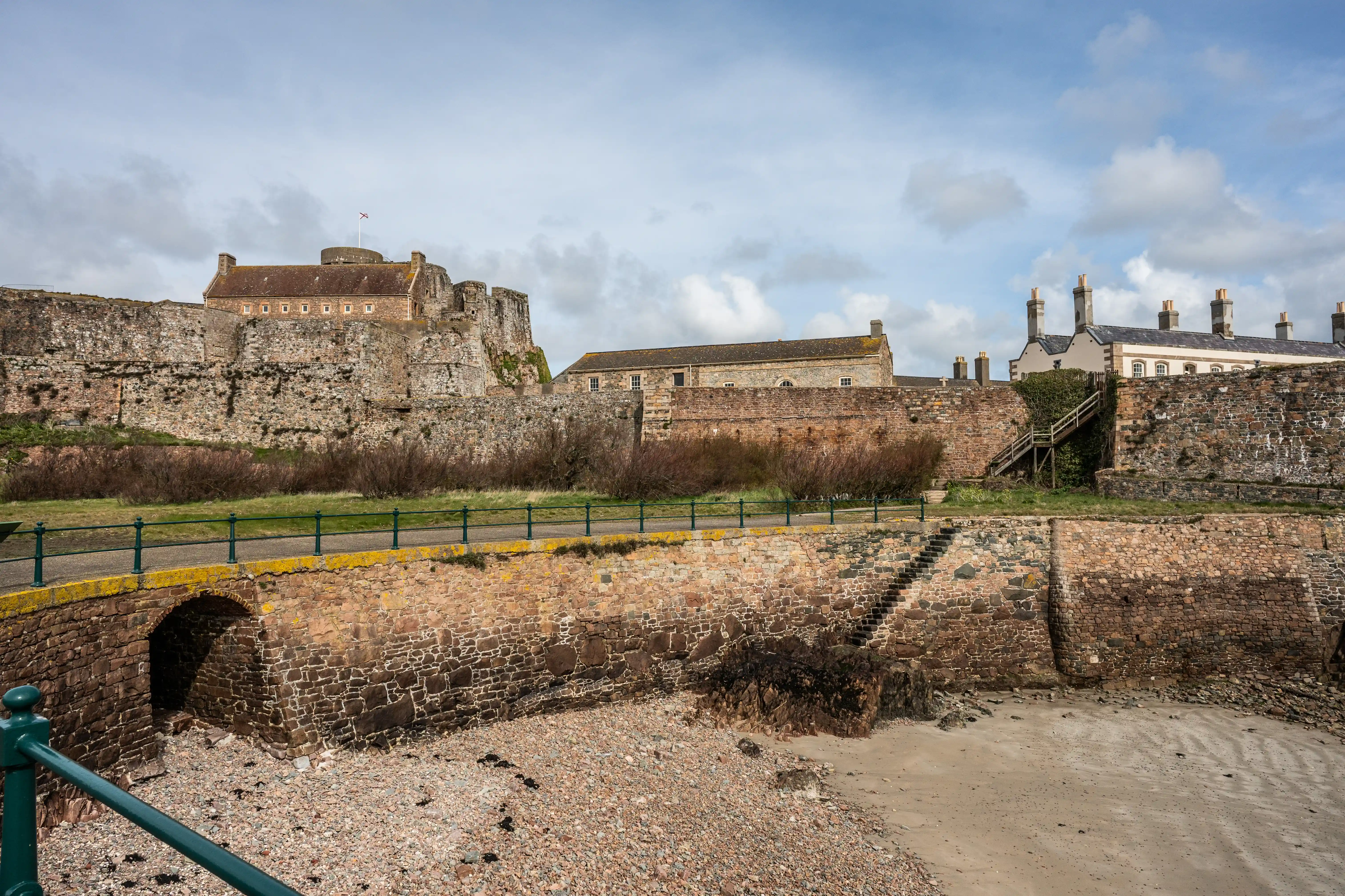 A stone castle with a red-tiled roof and battlements stands on a slight incline, with modern buildings behind it, all enclosed by sturdy stone walls. A path with a green railing runs in front of the walls, leading to an arched stone structure. The foreground shows a beach with sand and pebbles.