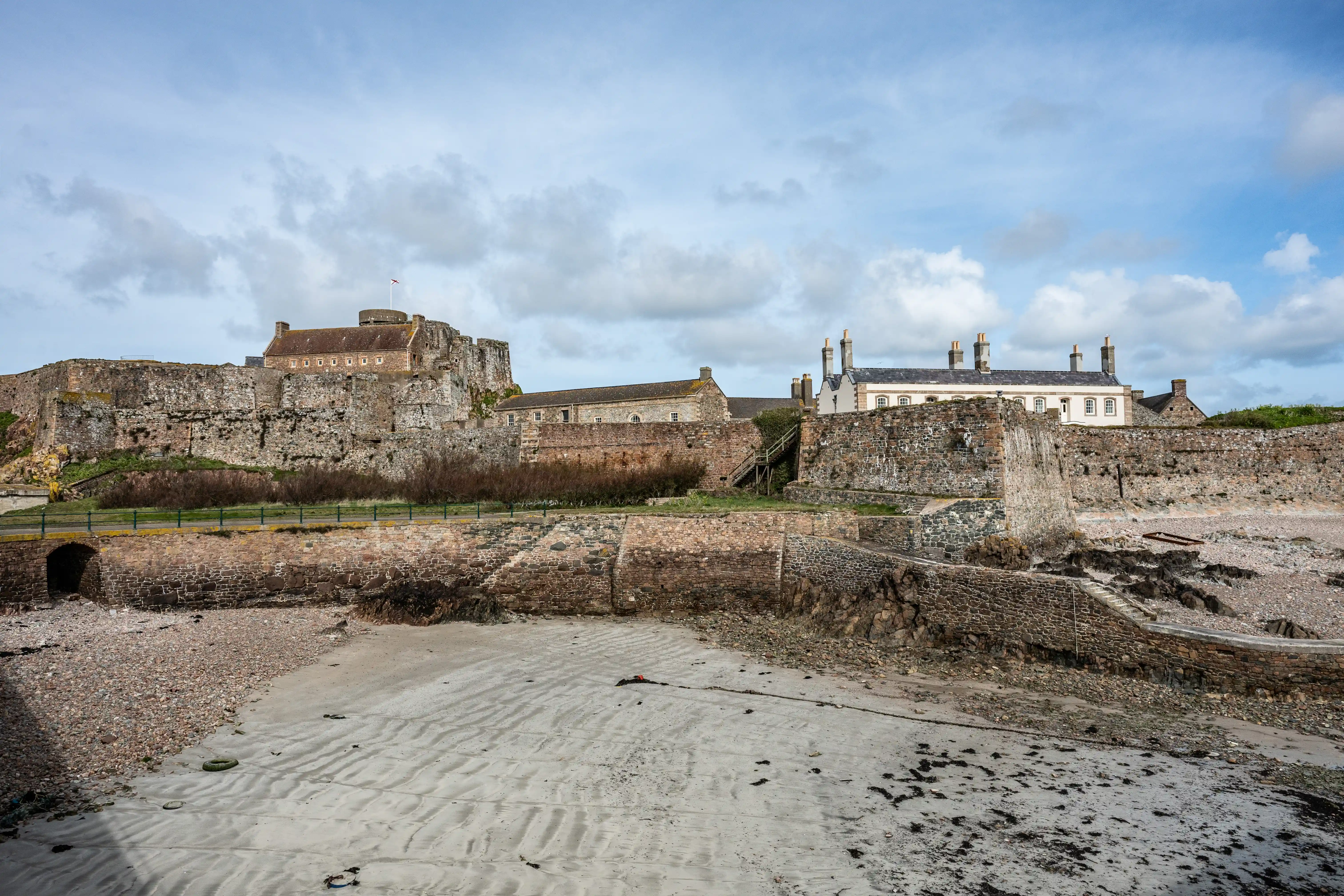 A wide shot of a large, ancient stone fortress with residential-style buildings attached, sitting above a beach. The sky is filled with grey and white clouds, and the foreground shows sand and pebbles.