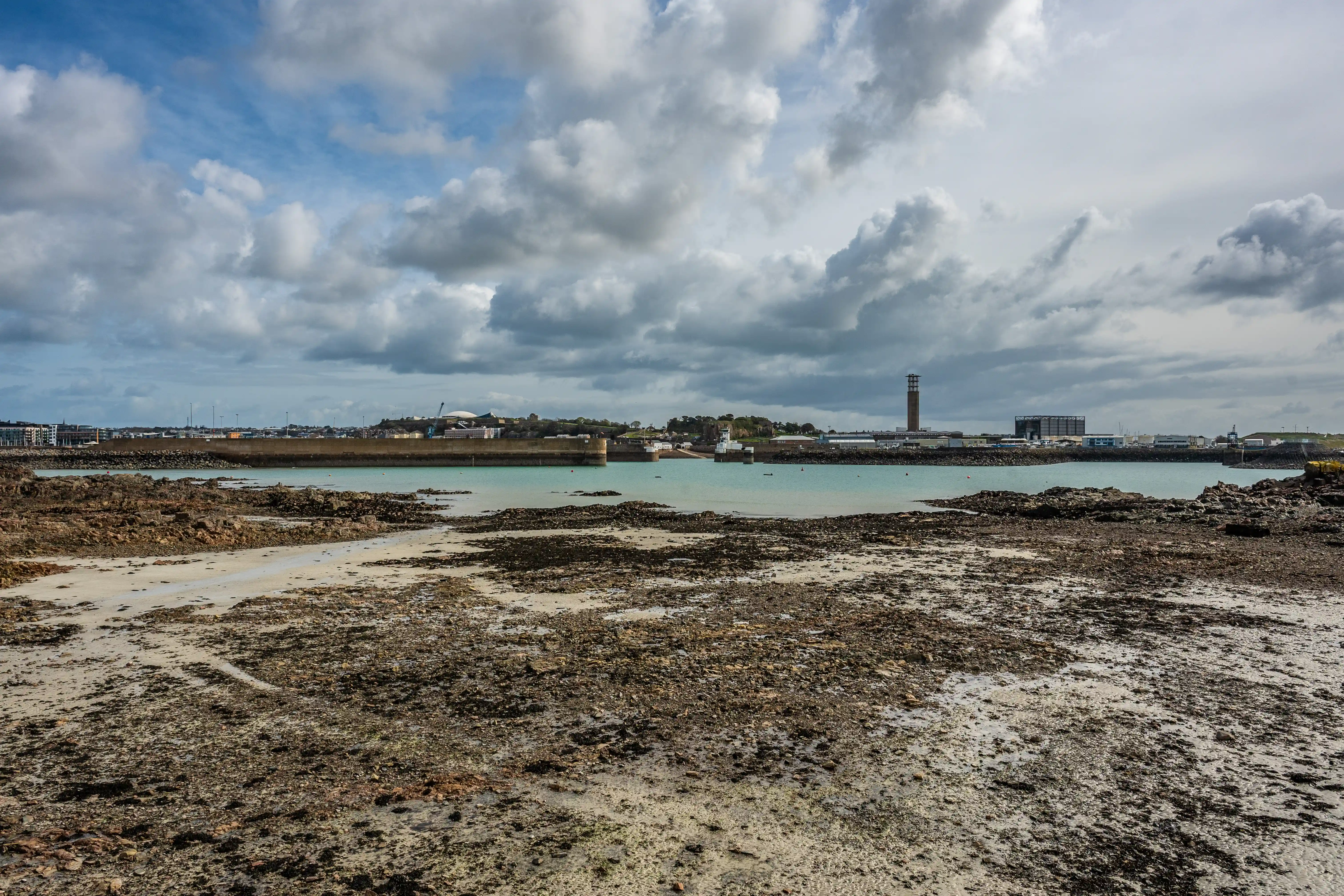 A view across a muddy, rocky shoreline at low tide towards a harbour with industrial buildings and a tall tower, under a cloudy sky.