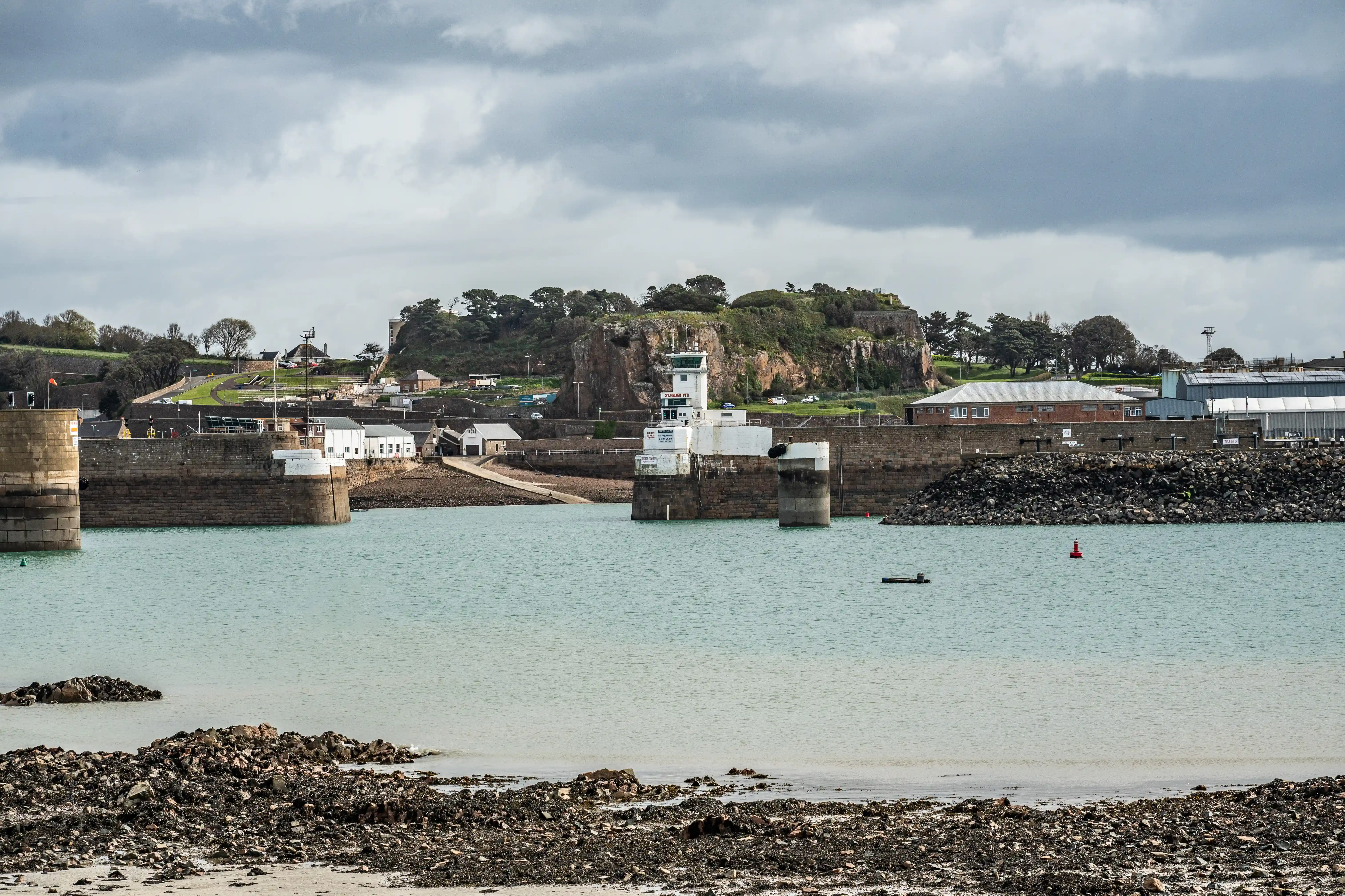 A scenic view of a harbour with a prominent white navigation tower, stone walls, and the sea. Rocky debris is visible in the foreground.