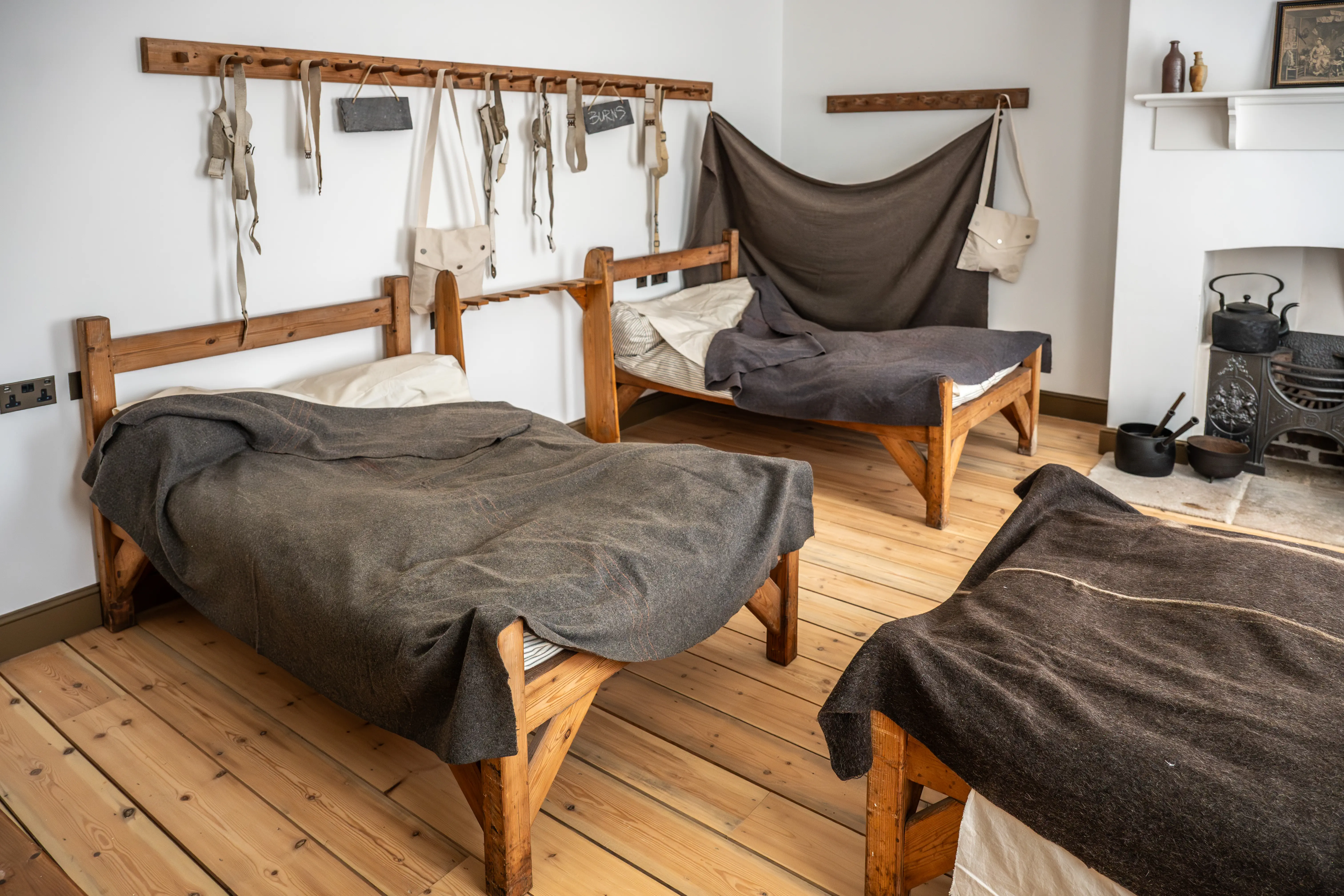 Three wooden beds with dark grey blankets and beige pillows in a rustic room. A rack with bags and straps hangs on the wall, and a fireplace is visible on the right.