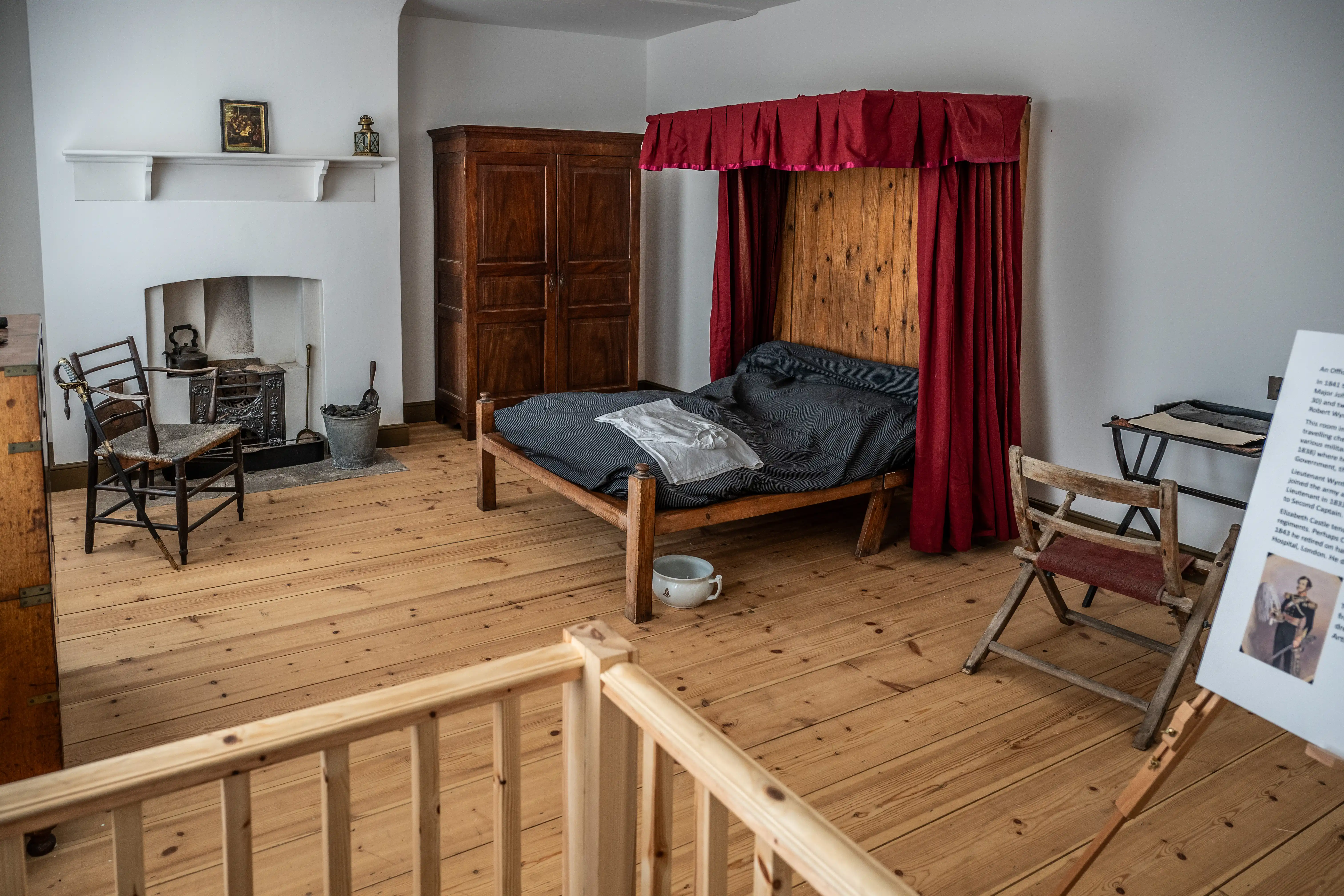 A historical room with a four-poster bed, wooden wardrobe, fireplace, and informational display, suggesting Victorian military quarters.