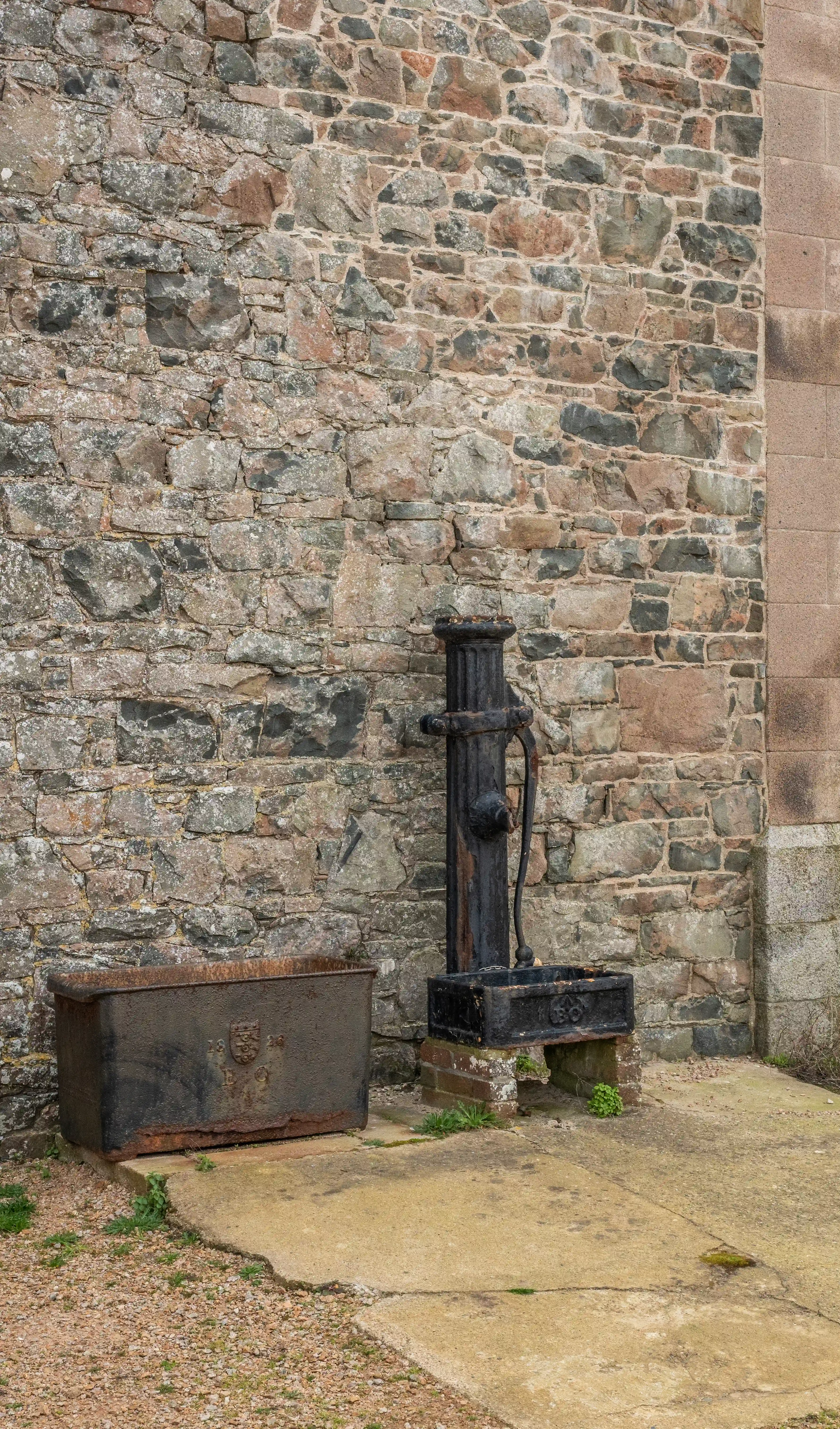 A black antique water pump and a rusty metal trough are positioned on a concrete surface in front of a rustic stone wall.