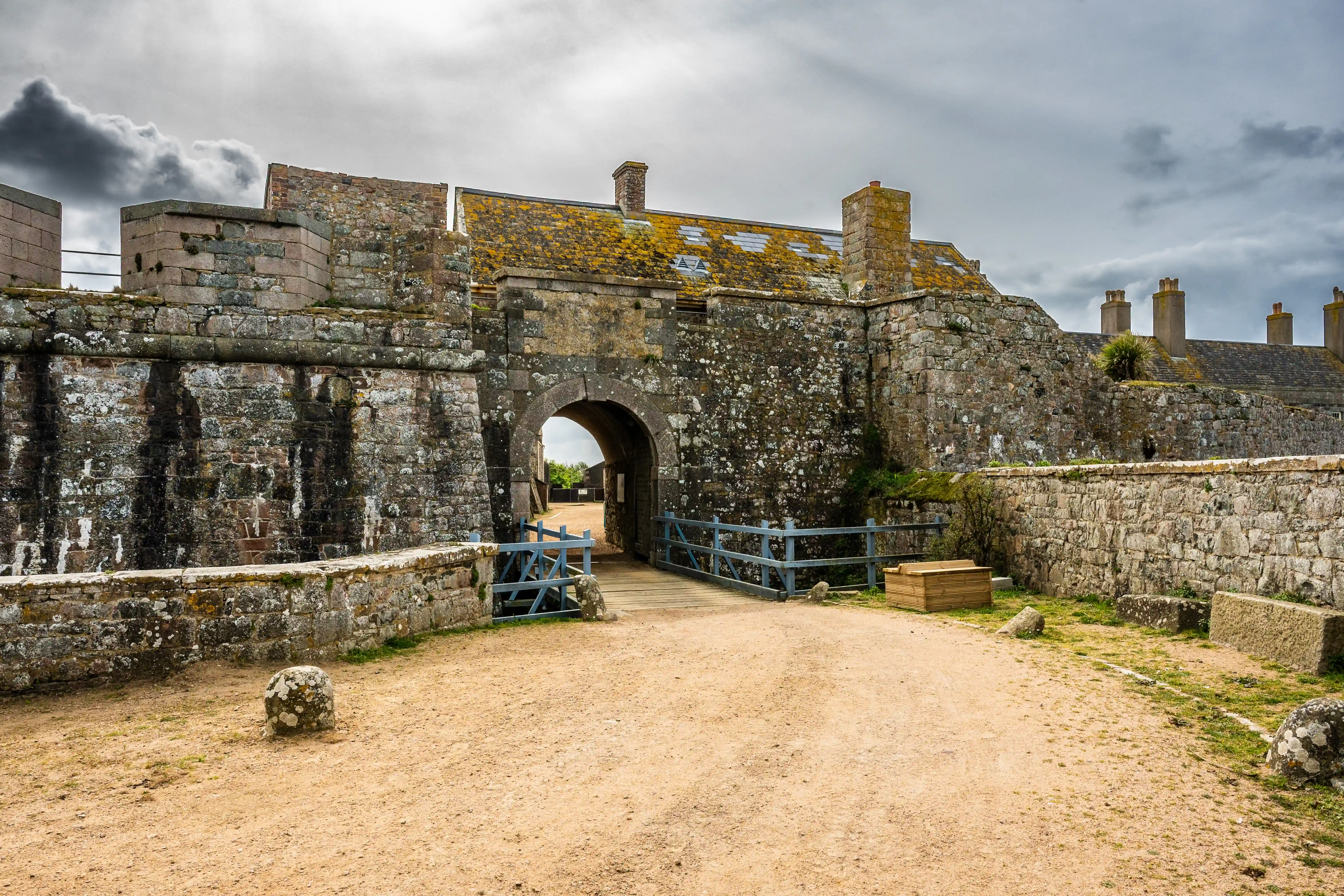 A photograph of a historical castle courtyard. In the foreground, there are several old stone buildings with dark grey slate roofs. Behind these buildings, a large, ancient stone castle is situated on a steep, grassy hill. A flag flies from the top of the castle. The ground in the courtyard is sandy earth.