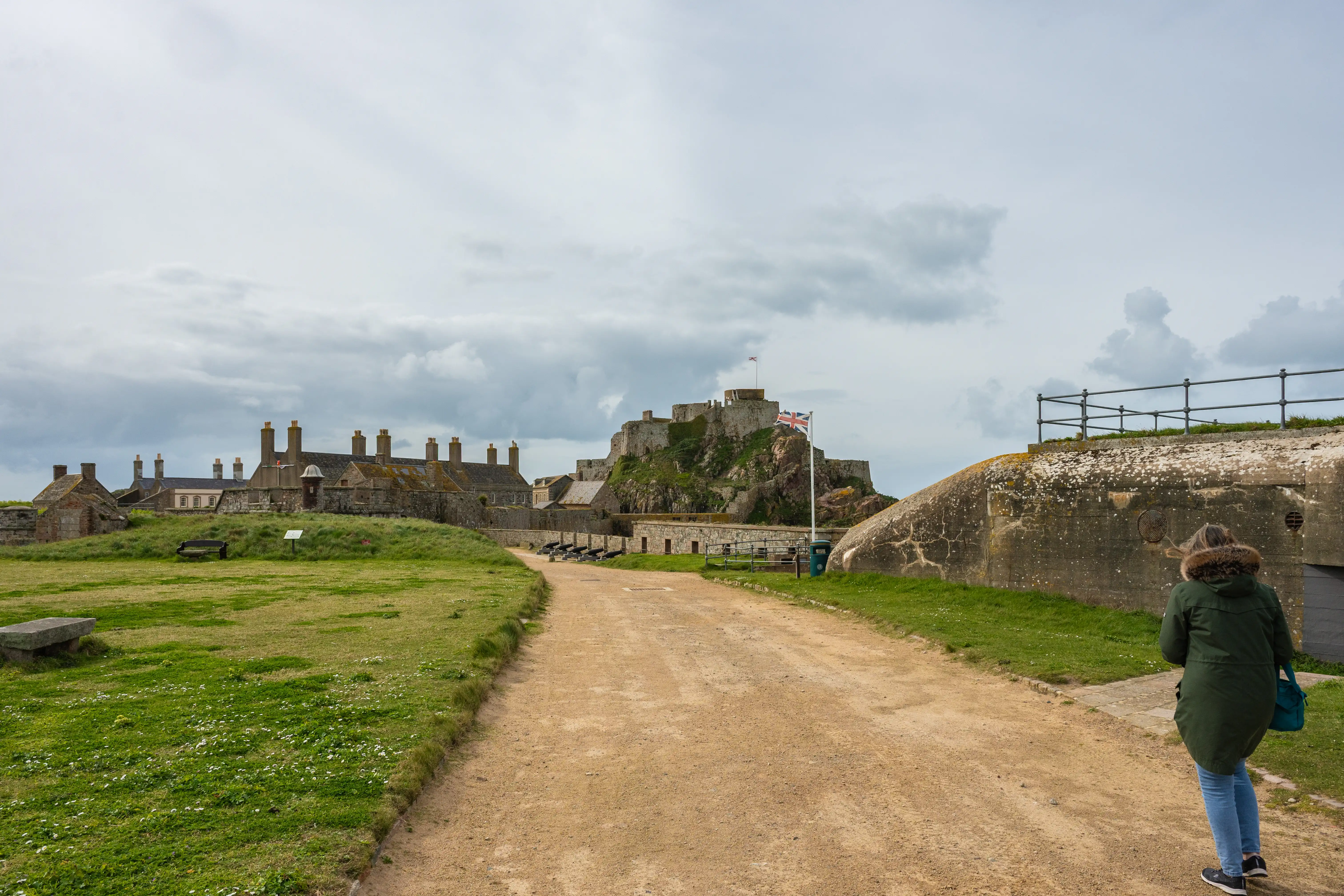 A person in a green coat walks down a dirt path towards a castle on a rocky hill, with a concrete fortification in the foreground and a cloudy sky overhead.