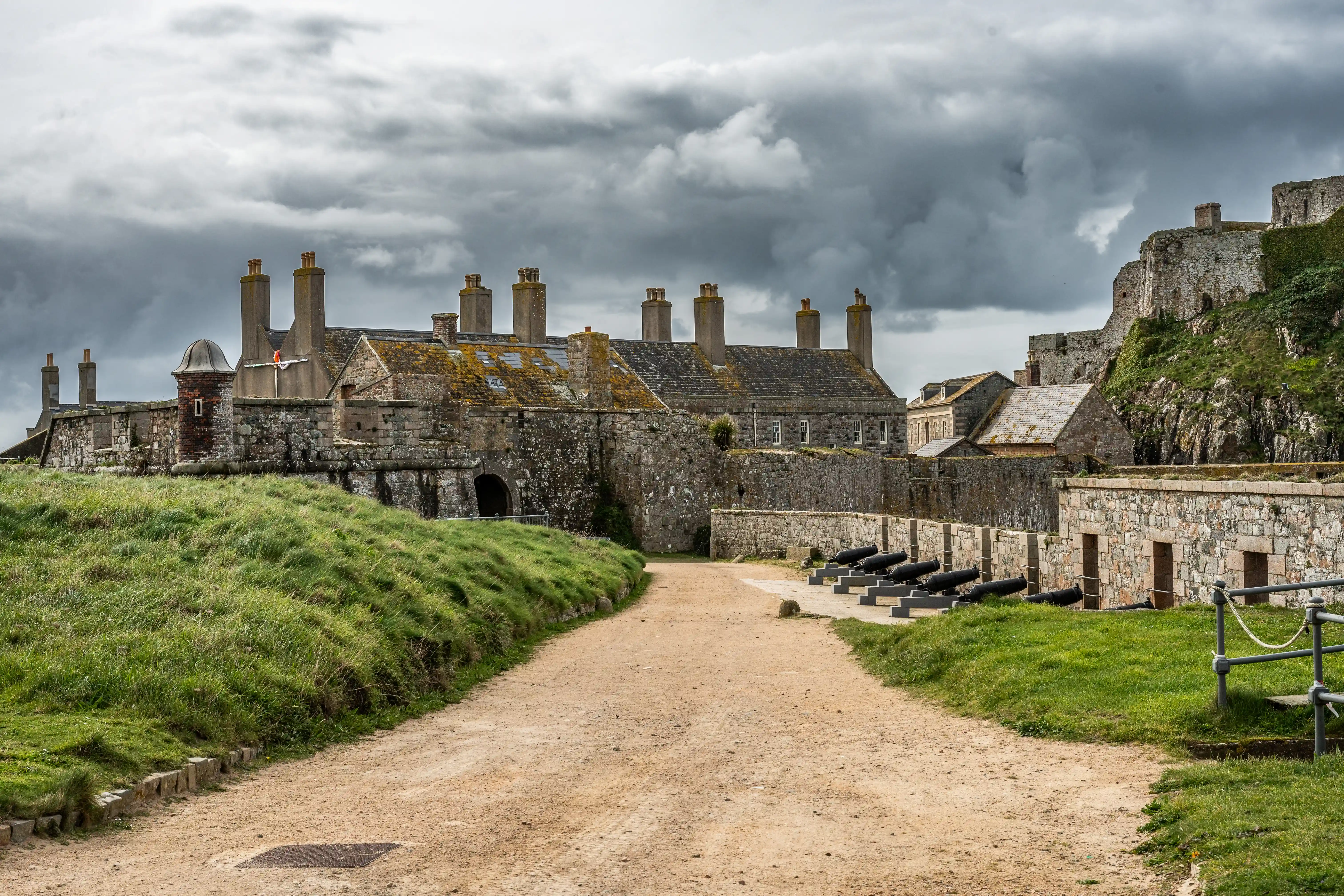 A gravel path leads up to a weathered stone fort with multiple chimneys and cannons lined up along a wall. The sky is overcast.