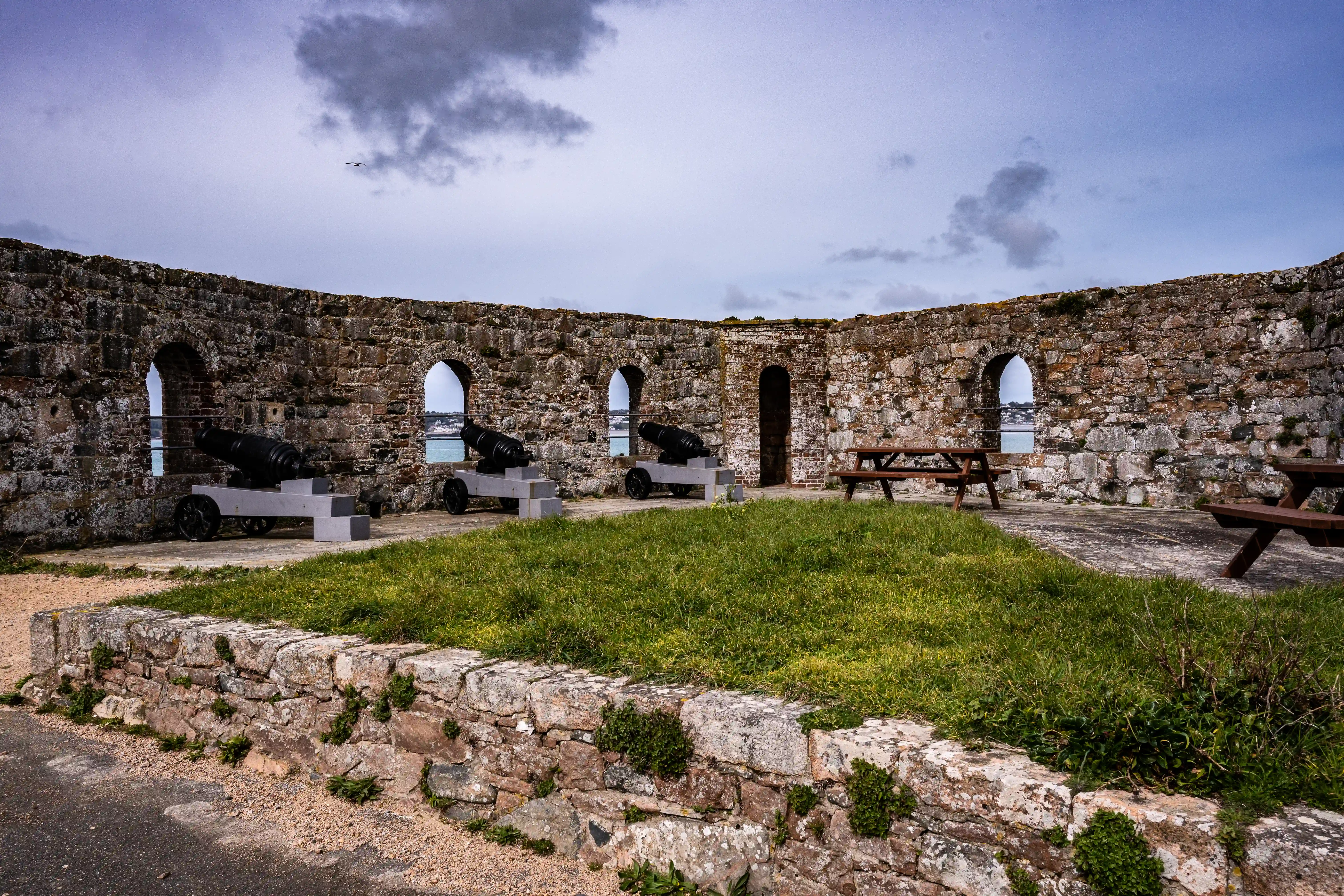 Stone fortification walls with arched openings revealing the sea, two black cannons on mounts, and wooden picnic tables.