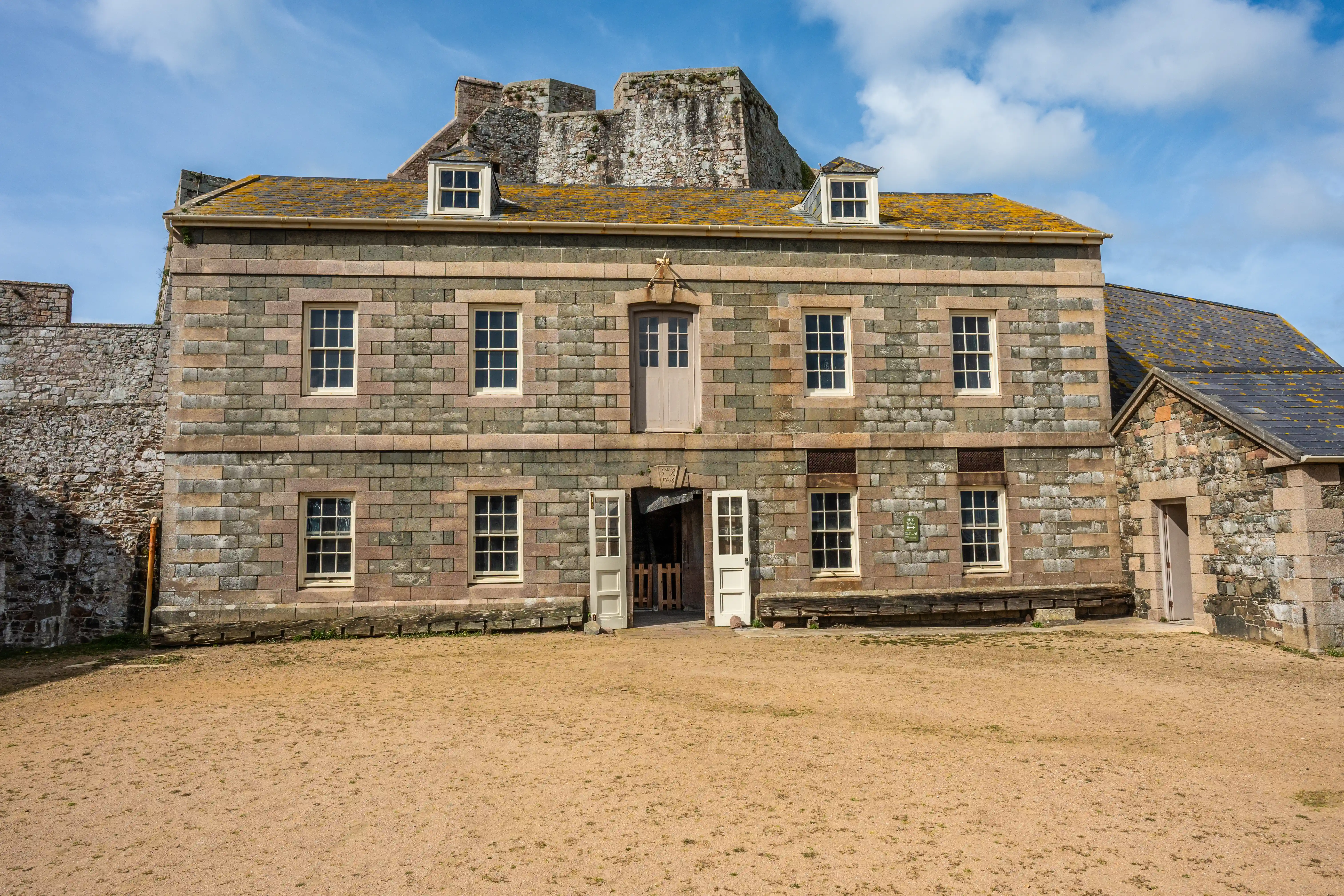 A two-storey stone building with multiple sash windows and an open central doorway, situated in front of a stone fortification under a blue sky with clouds.