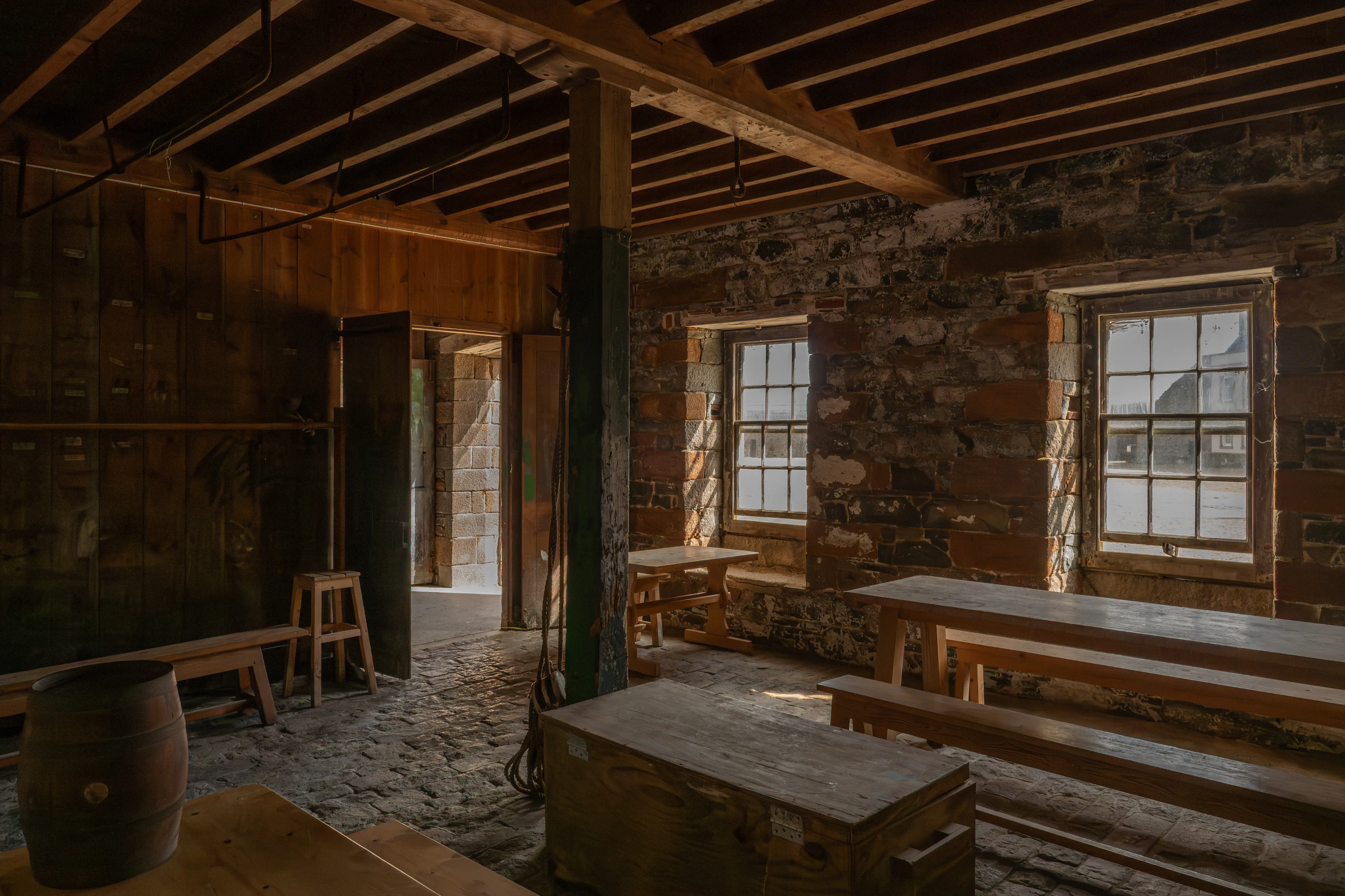 Interior of a rustic building with rough stone walls, wooden beamed ceiling, cobbled floor, wooden tables, benches, a barrel, and doorways.